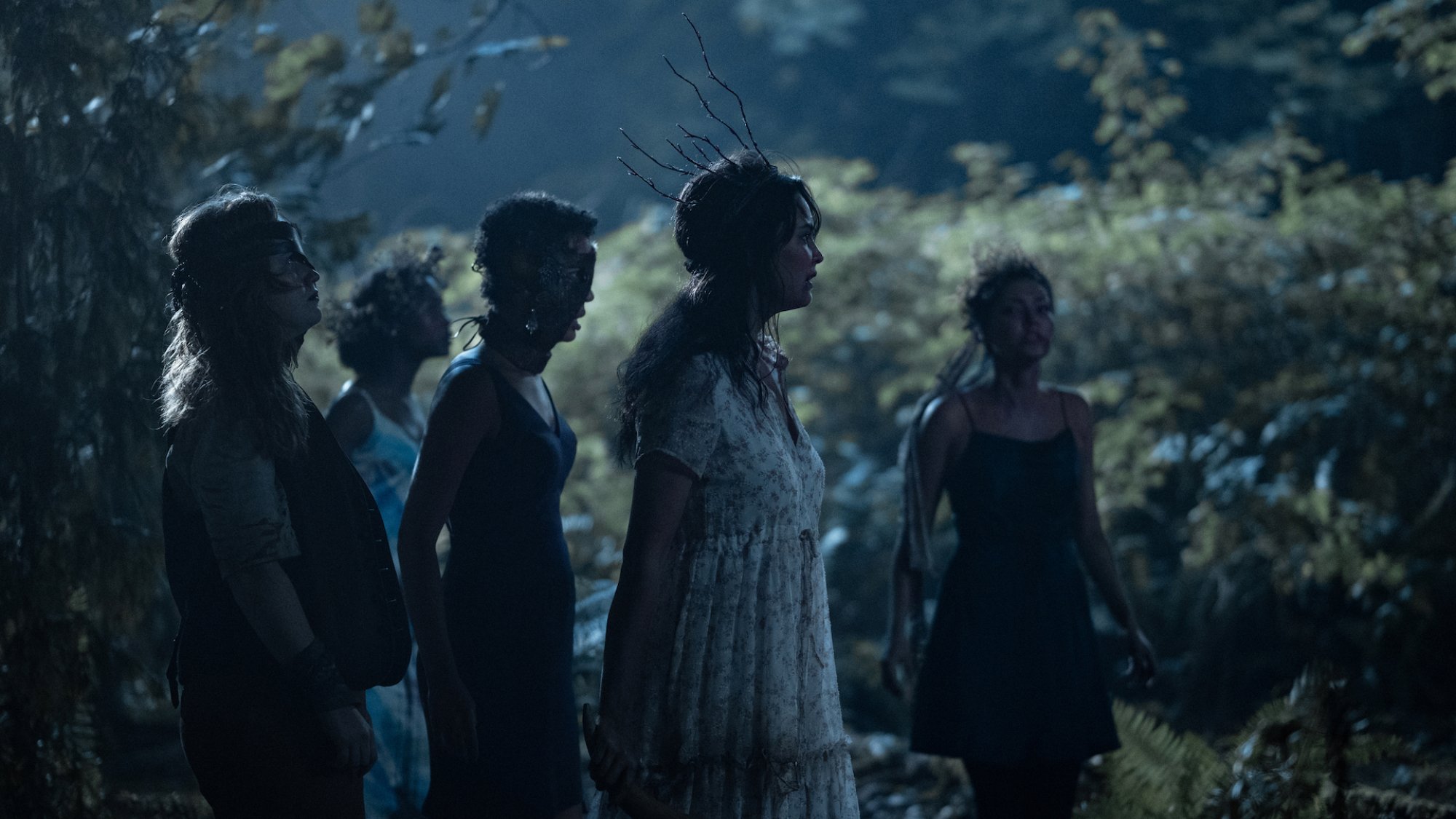 Un groupe de filles portant des couronnes de brindilles se tient dans un bois dans l'obscurité.