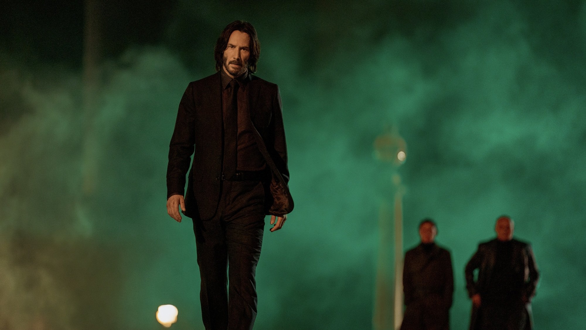 Un homme en costume noir traverse une fumée verte.  Deux silhouettes sombres marchent loin derrière lui.