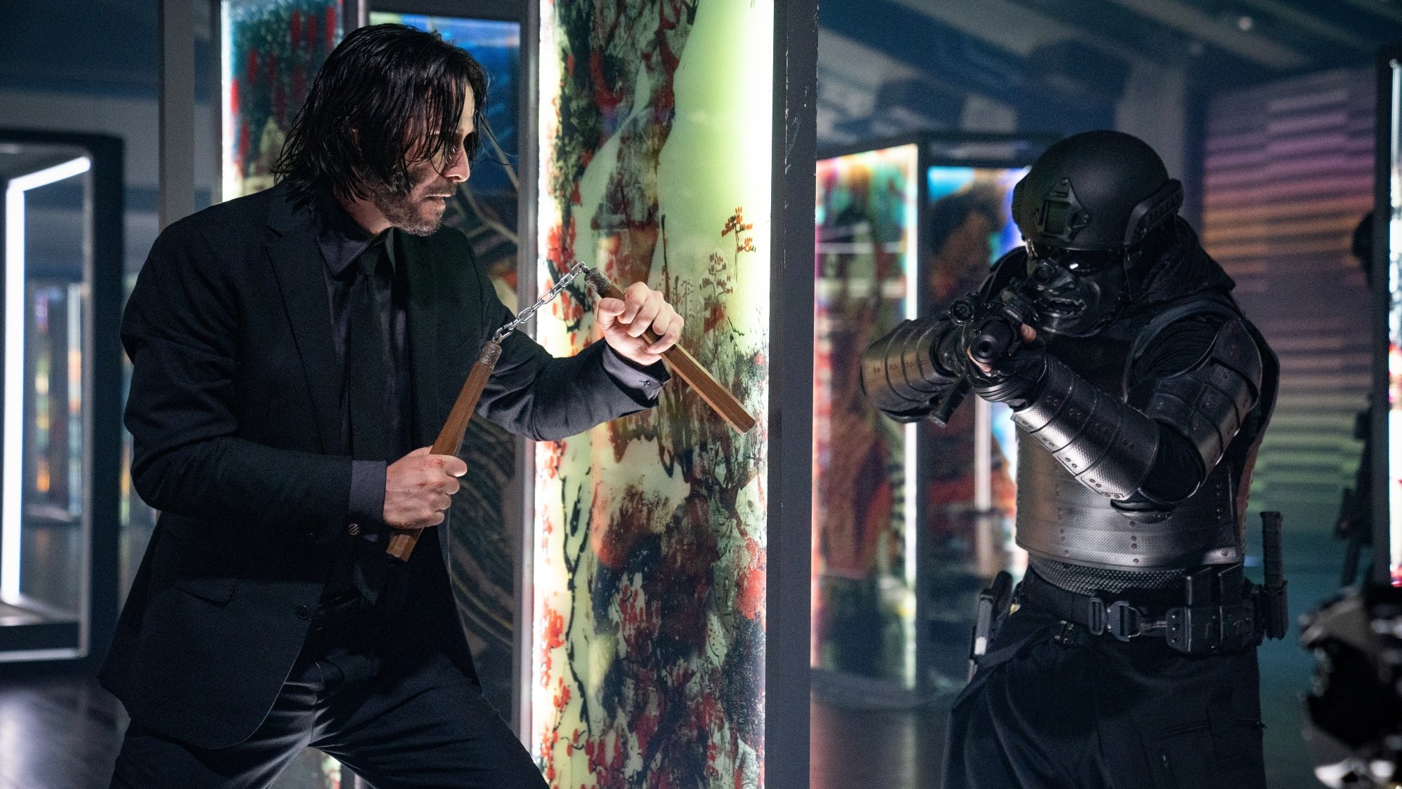 Un homme en costume noir brandit des nunchuks contre un homme armé portant une armure tactique noire.