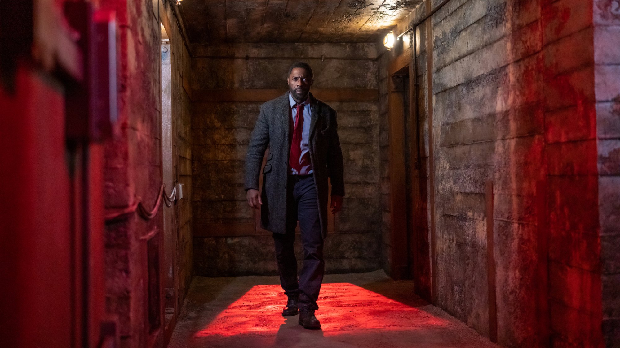 Un homme en costume et trench-coat marche à grands pas dans un couloir industriel éclairé en rouge.