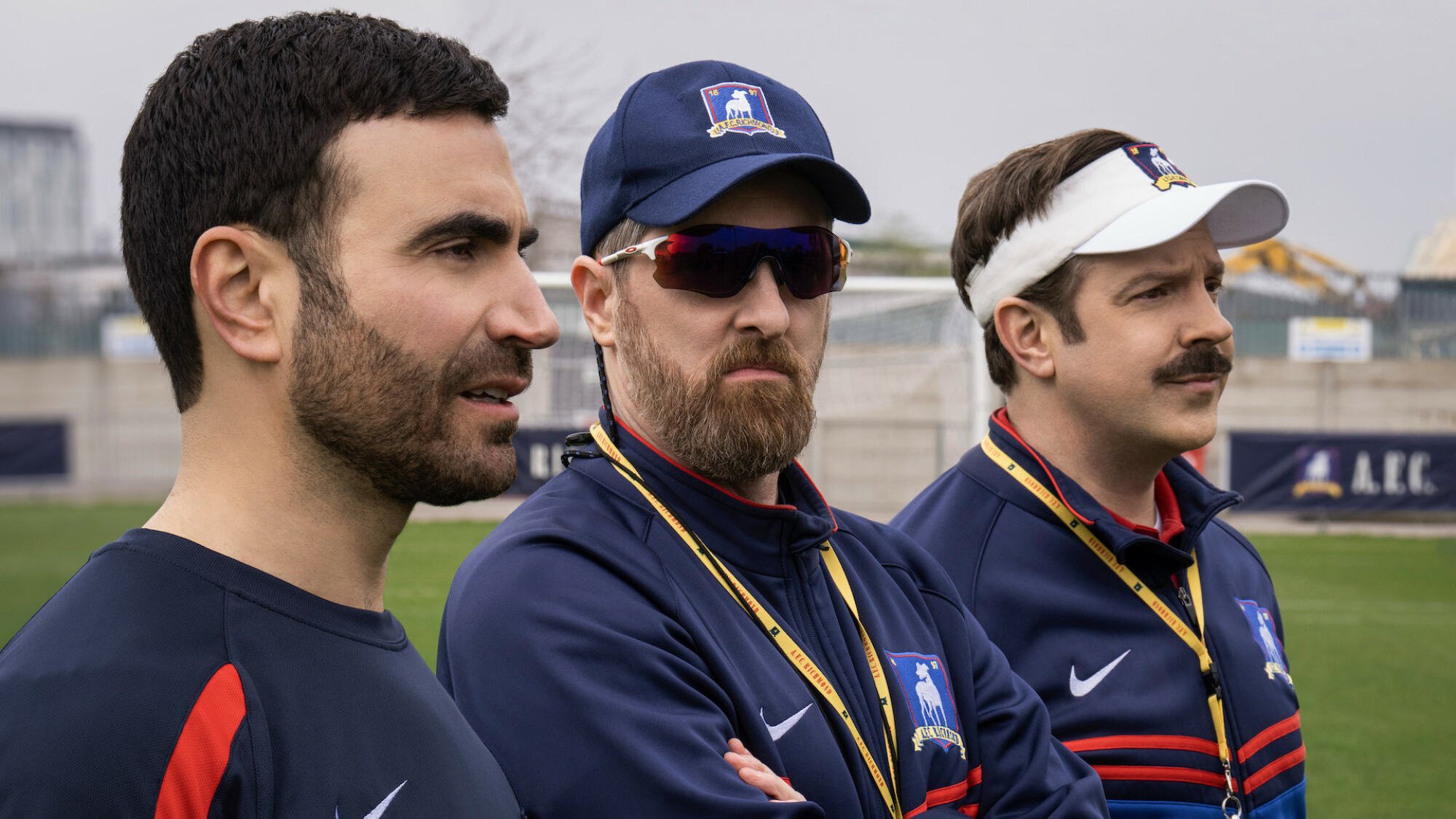 Un joueur de football et deux entraîneurs de football se tiennent sur le côté d'un terrain pendant l'entraînement.