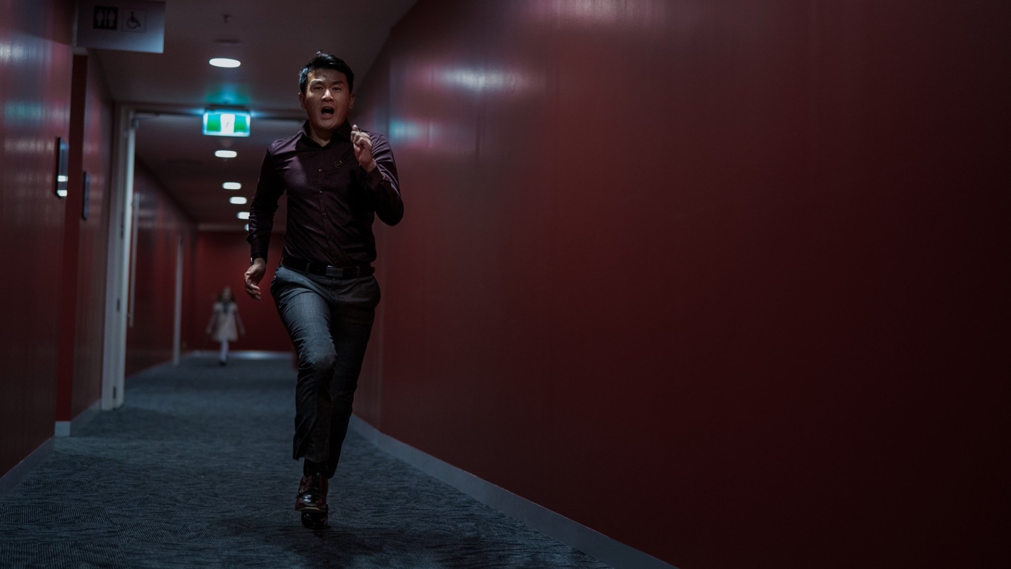 Un homme court dans un couloir aux murs rouge foncé tandis qu'une poupée grandeur nature se cache en arrière-plan.