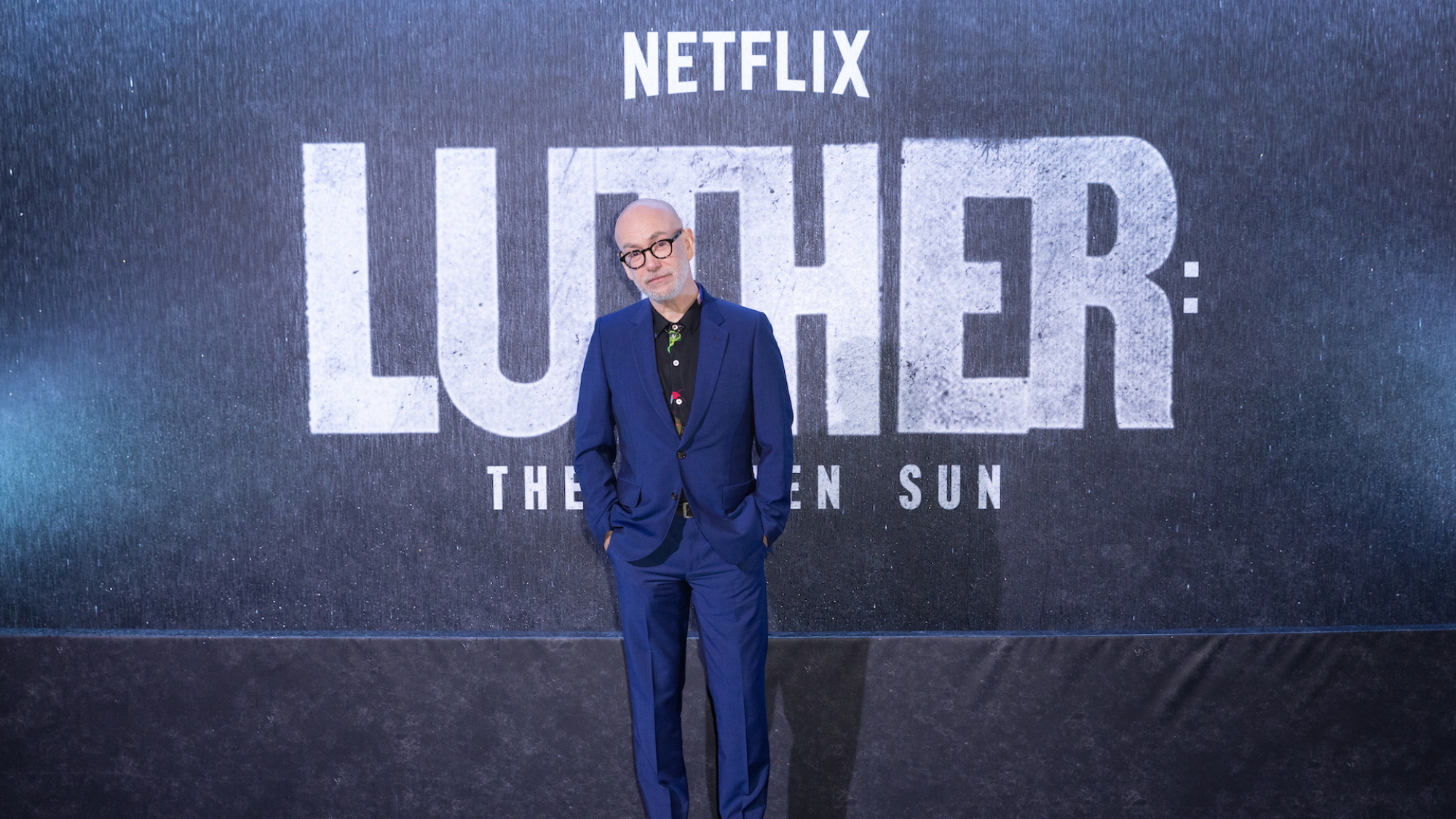 Un homme en costume bleu et lunettes à monture épaisse se tient à une première de film avec les mots 