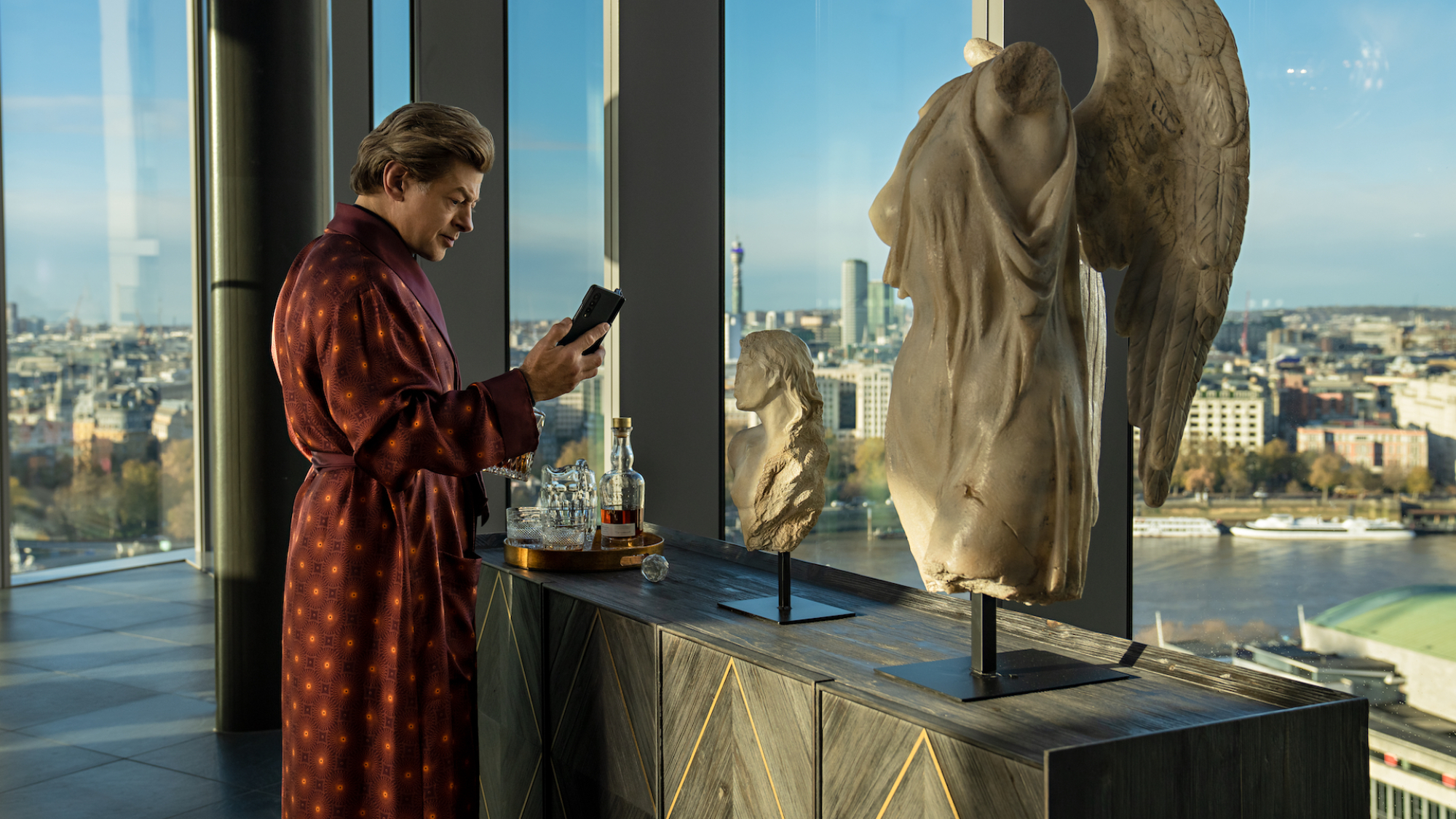 Un homme riche en peignoir utilise un téléphone pliable dans un appartement entièrement vitré près d'un buste en marbre.