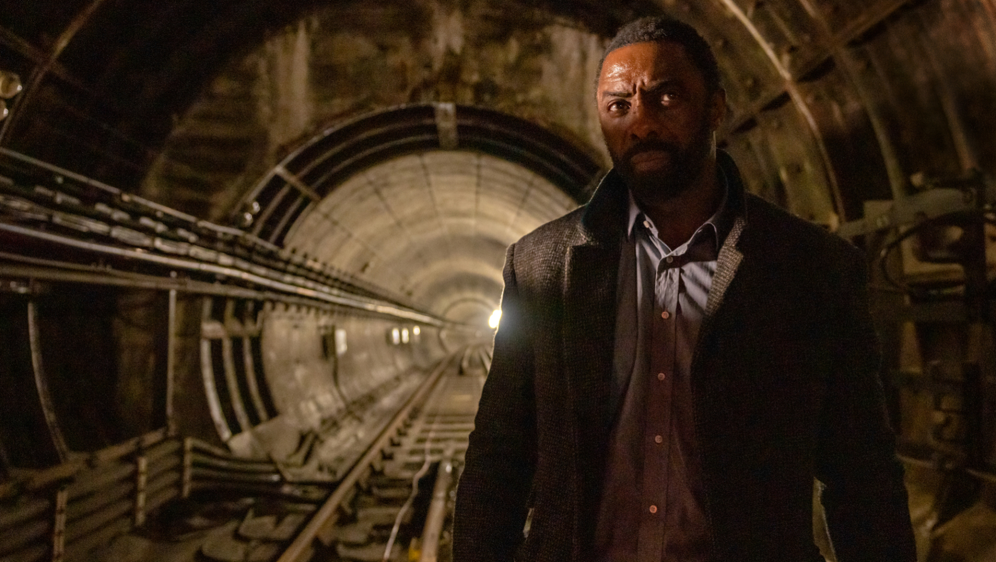 Un homme en costume et trench-coat se tient dans un tunnel vide du métro de Londres avec la lumière mettant en valeur ses yeux.