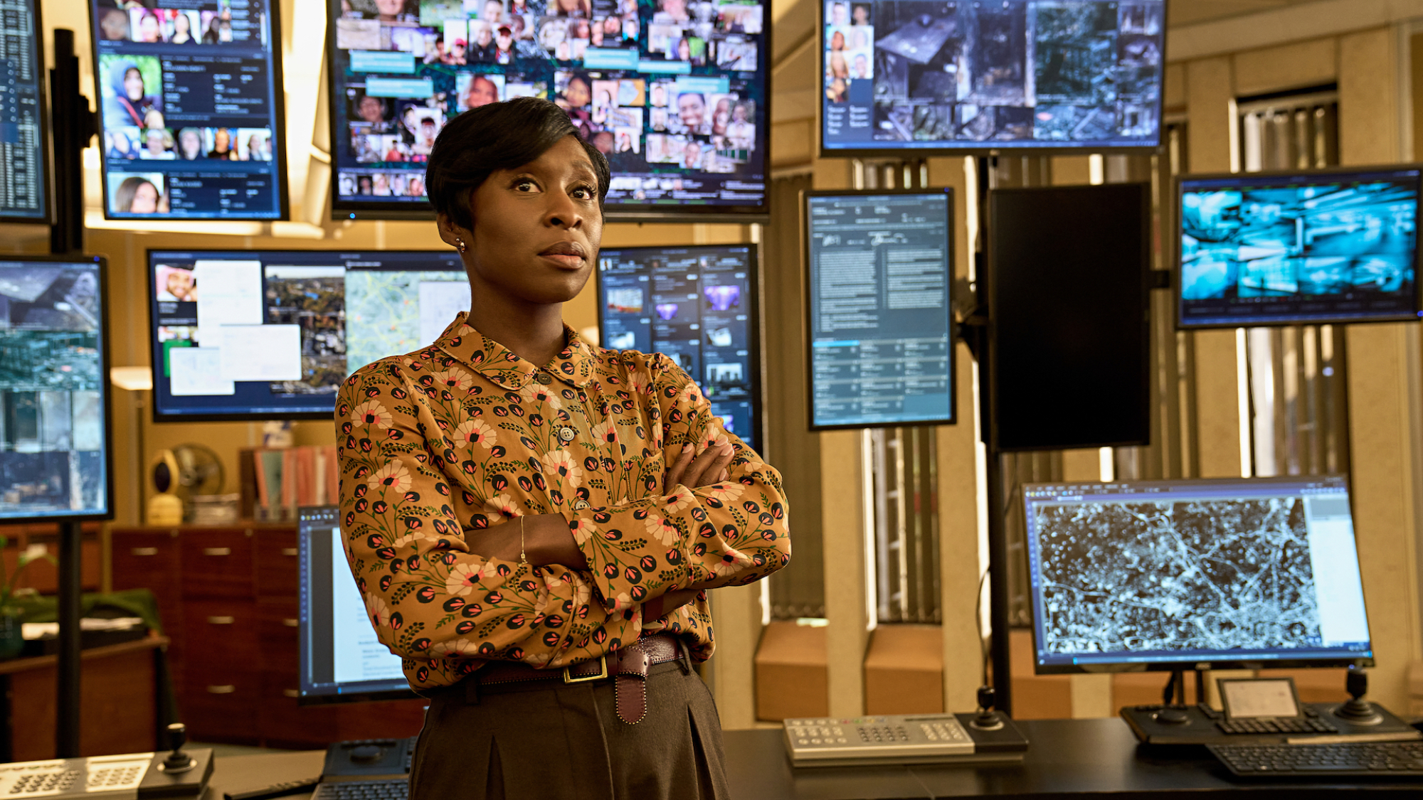 Une femme portant une chemise à motifs se tient dans une salle de surveillance de la police avec environ 10 écrans derrière elle montrant diverses images et informations de suivi.