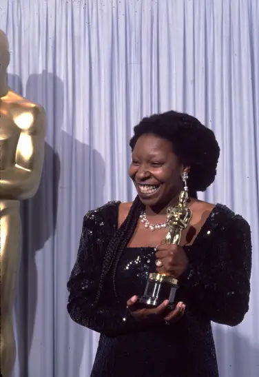 Whoopi Goldberg brandit un Oscar devant un fond blanc. 