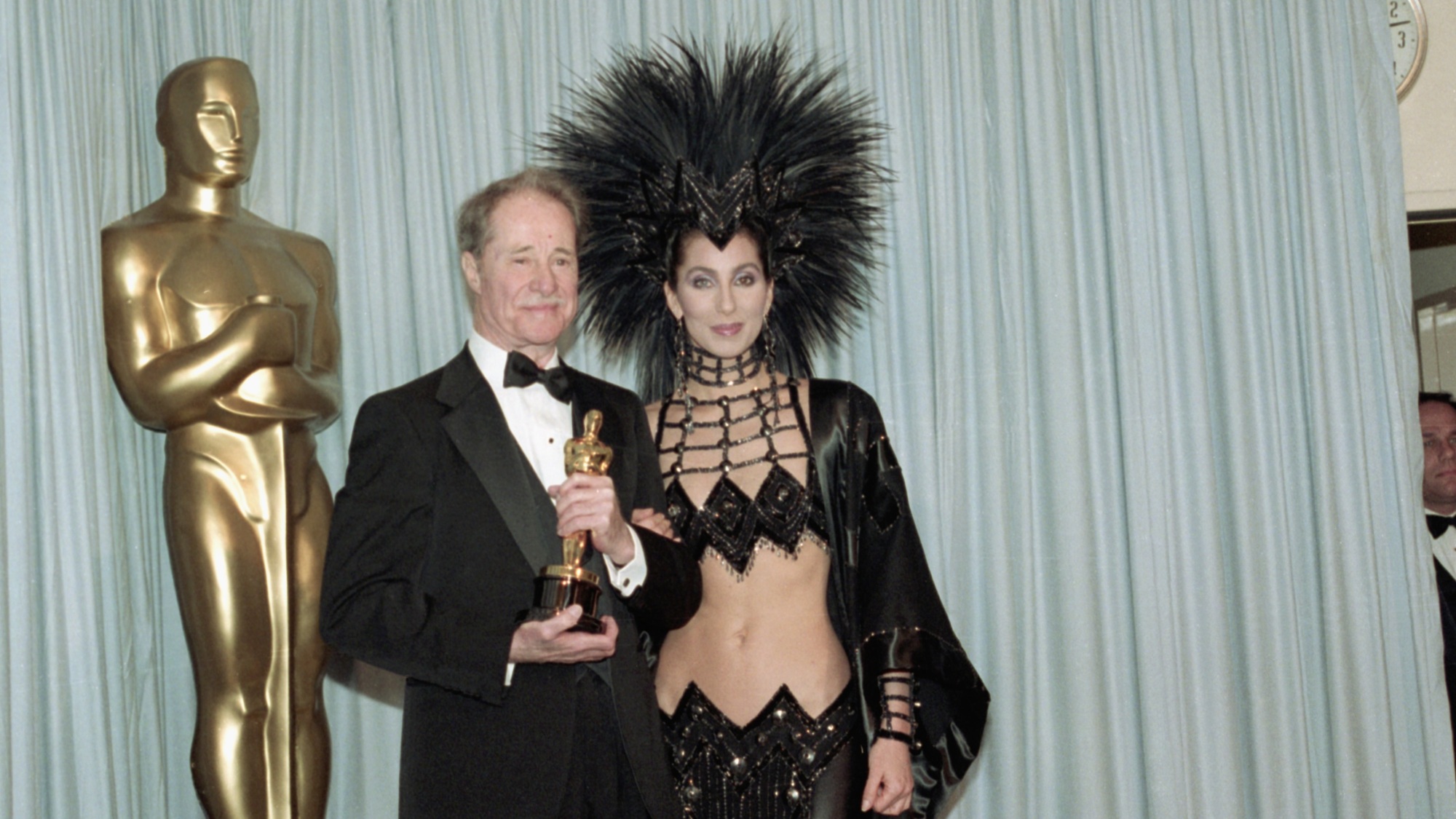 Cher dans une robe noire en deux pièces avec une coiffe massive à paillettes scintillantes.  Elle se tient à côté de l'acteur Don Ameche, qui tient un Oscar.