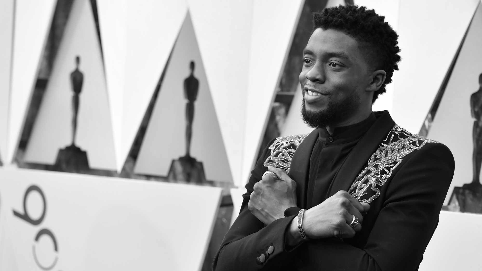 L'acteur Chadwick Boseman en costume noir sur le tapis rouge des Oscars.  Il croise les bras sur sa poitrine lors du salut Wakanda.