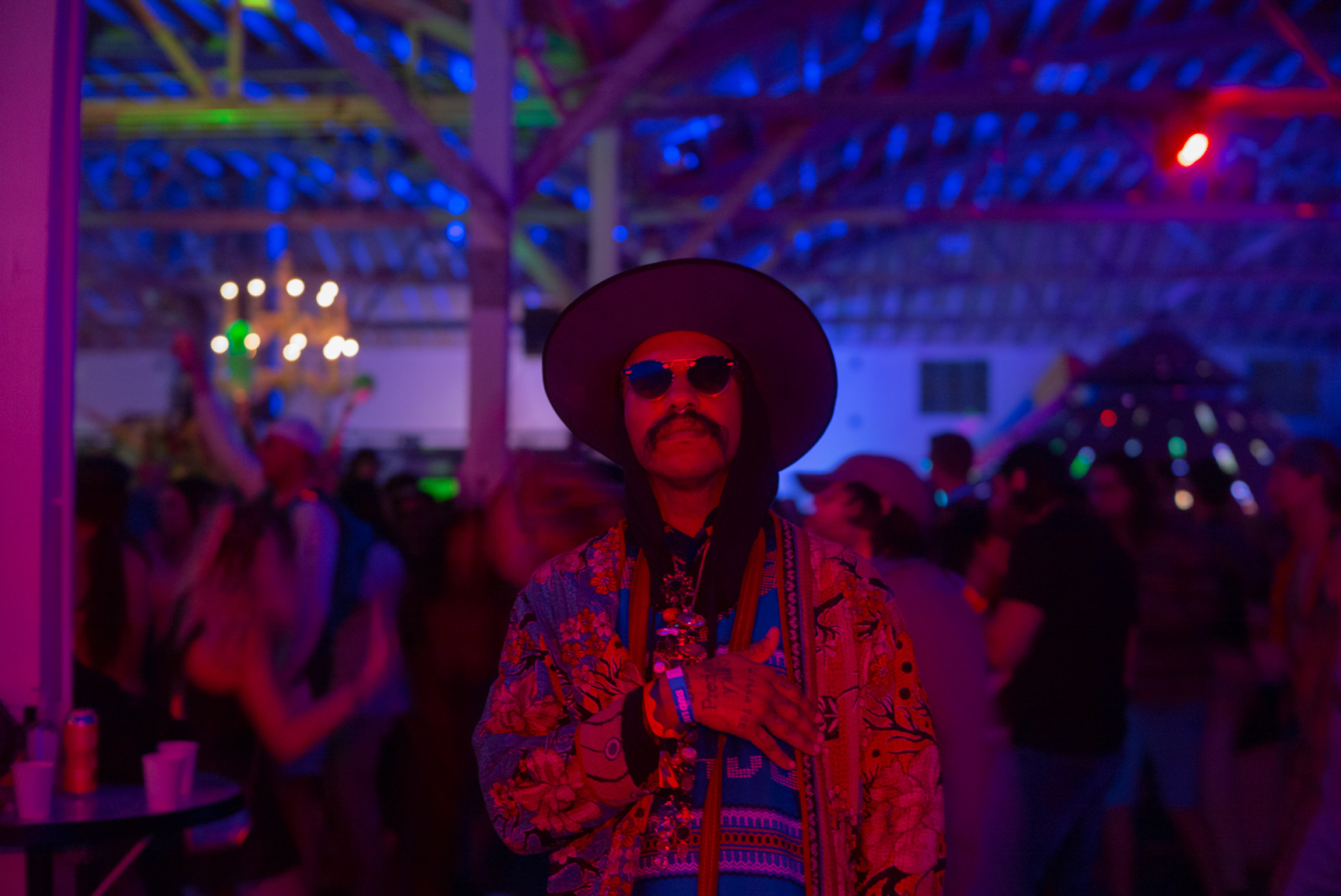 Un festivalier pose sous une lumière rouge et bleue avec une foule dansante derrière lui.
