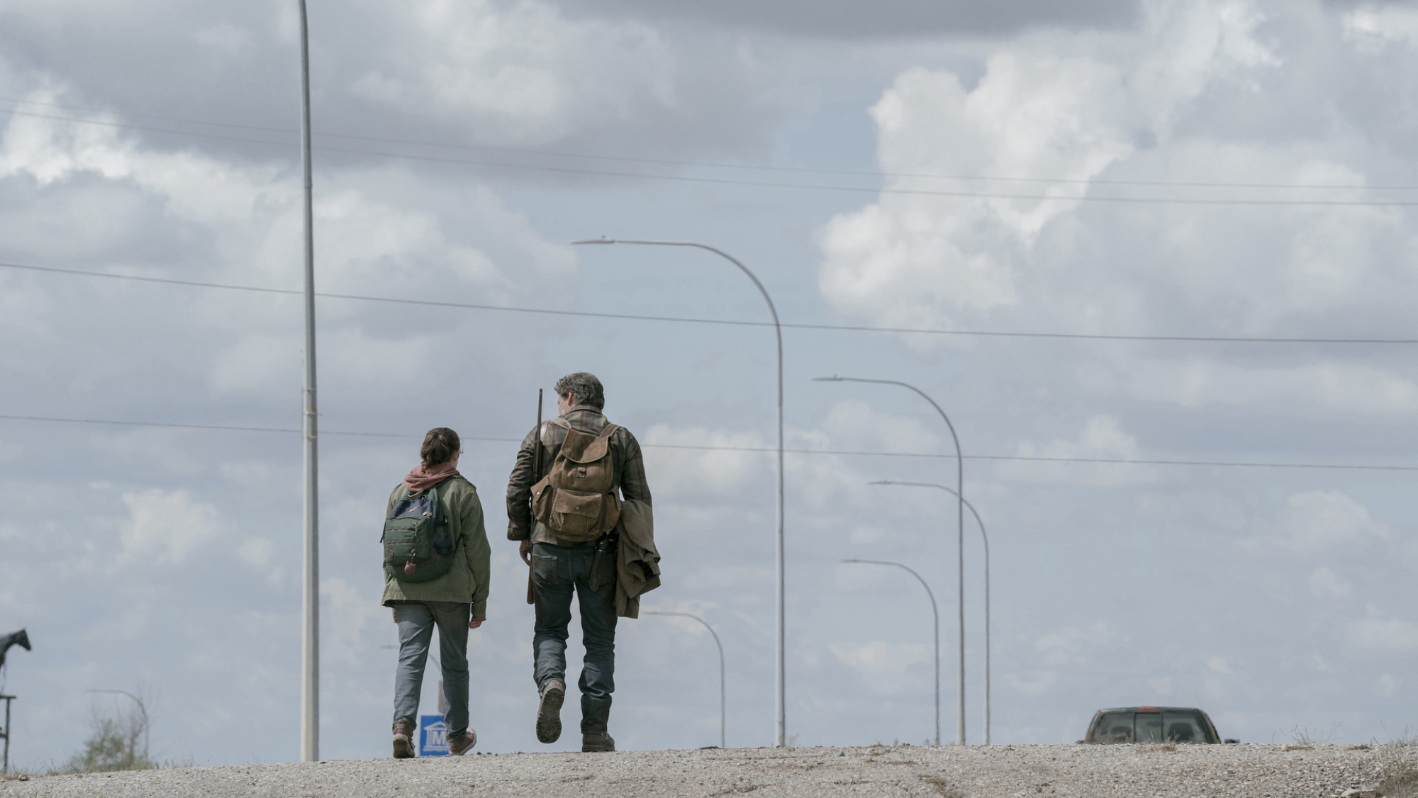 Un homme et un adolescent portant des sacs à dos et des armes à feu marchent sur une route abandonnée.