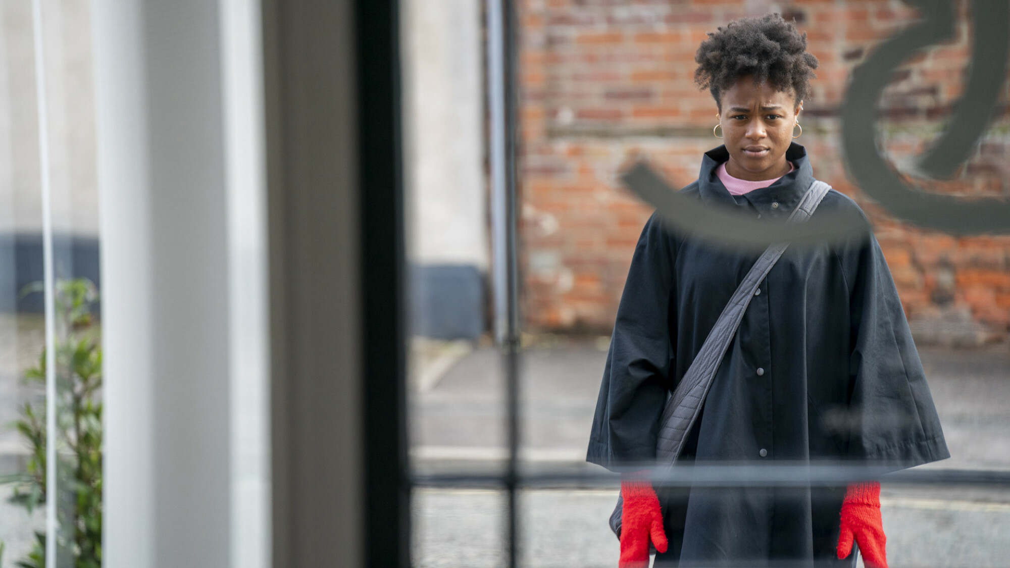 Une jeune femme se tient à l'extérieur, vue à travers une fenêtre.  Elle fait face à la caméra et porte des gants rouge vif.