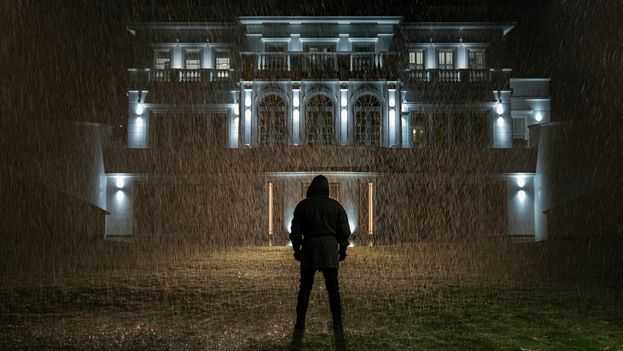 Un homme cagoulé se tient sous la pluie et regarde fixement une grande maison.