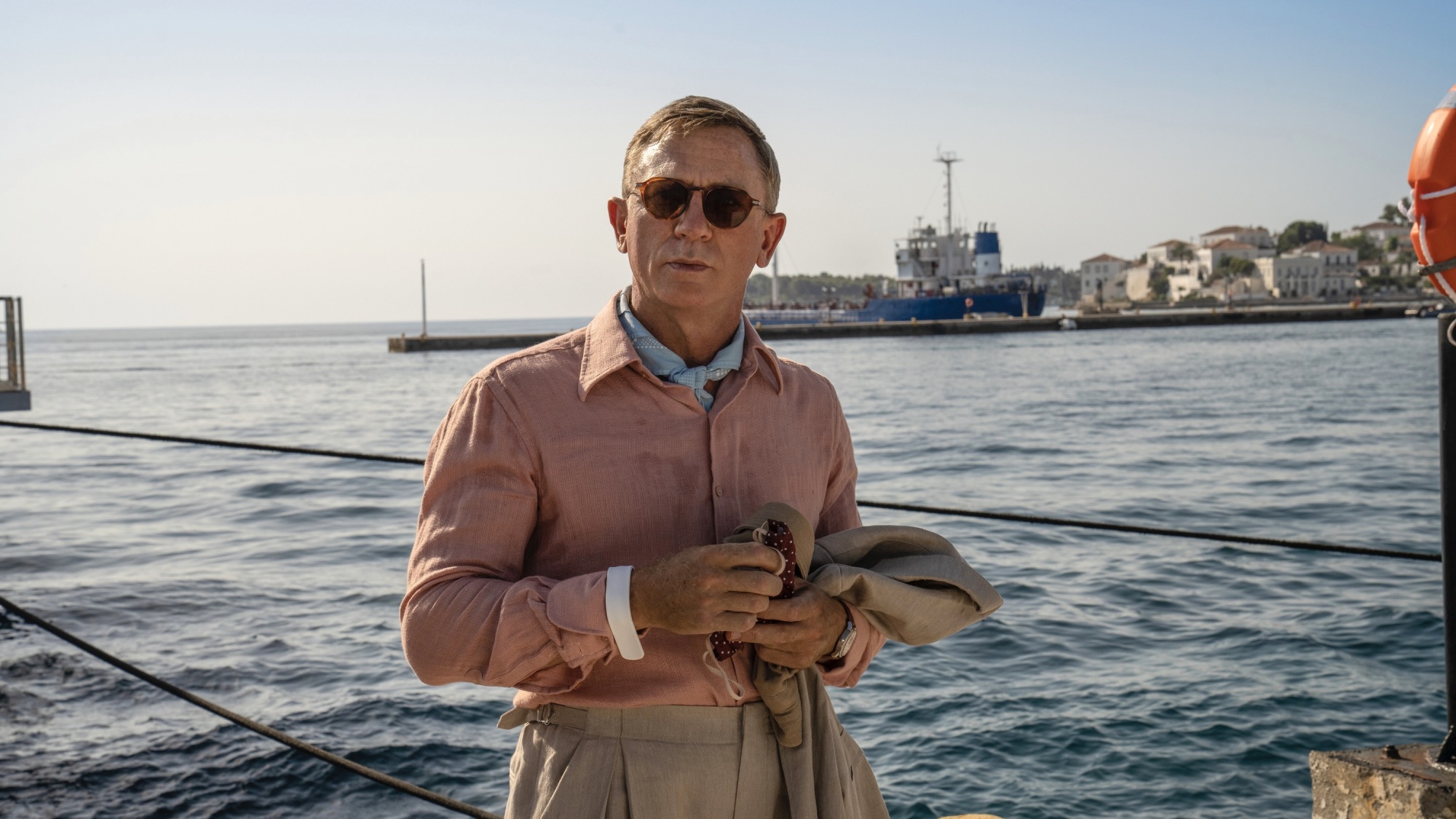 Daniel Craig, un homme blanc vêtu d'une chemise en lin rose et de lunettes de soleil, se tient sur un quai au bord de l'océan.