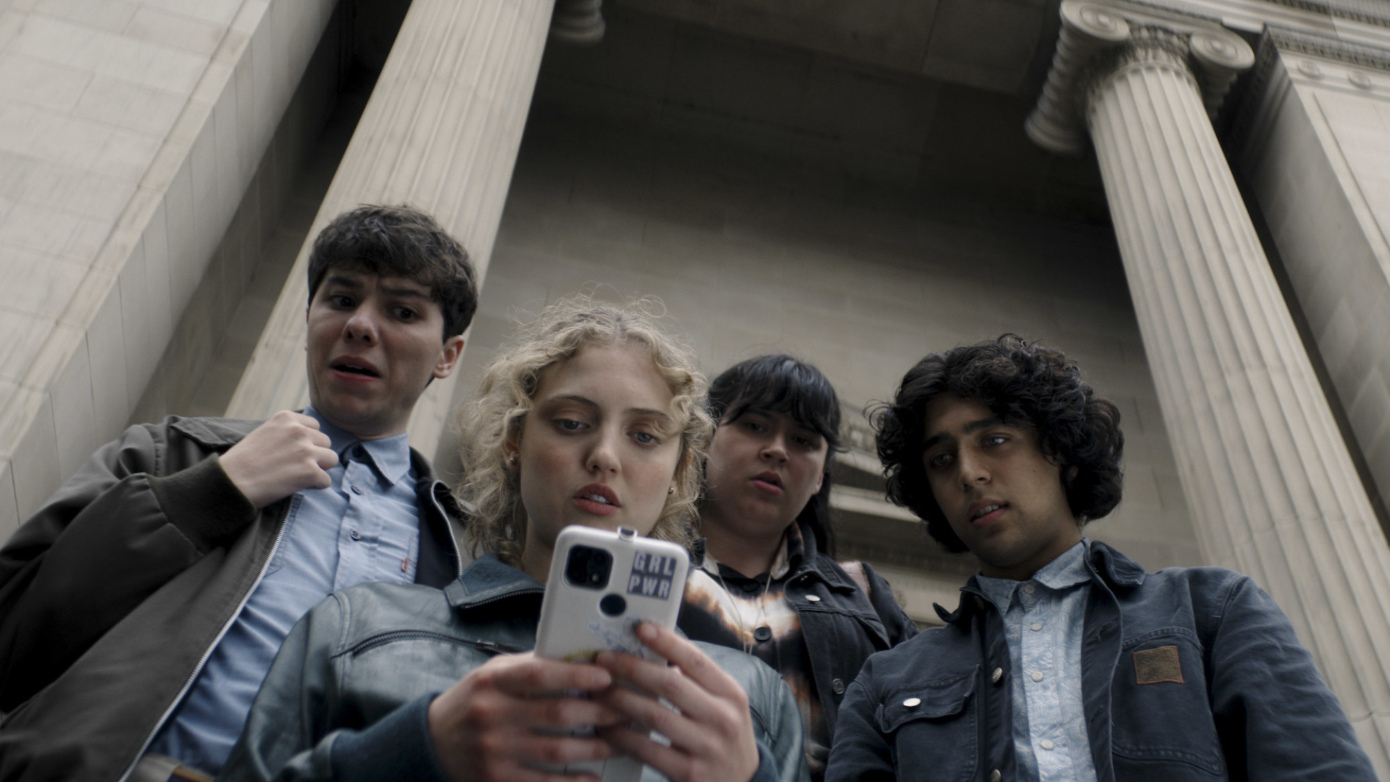 Un groupe d'adolescents se rassemble devant les colonnes d'un bâtiment gouvernemental, autour d'une adolescente utilisant son téléphone.
