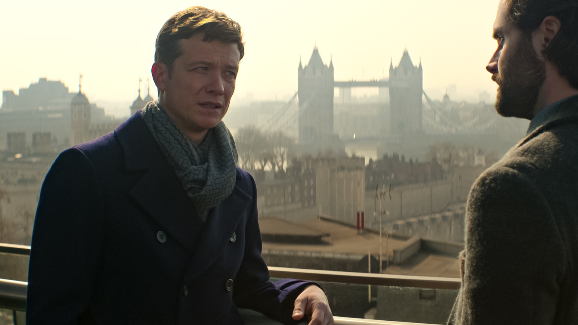 Un homme en trench-coat parle à son ami sur un balcon surplombant Londres. 