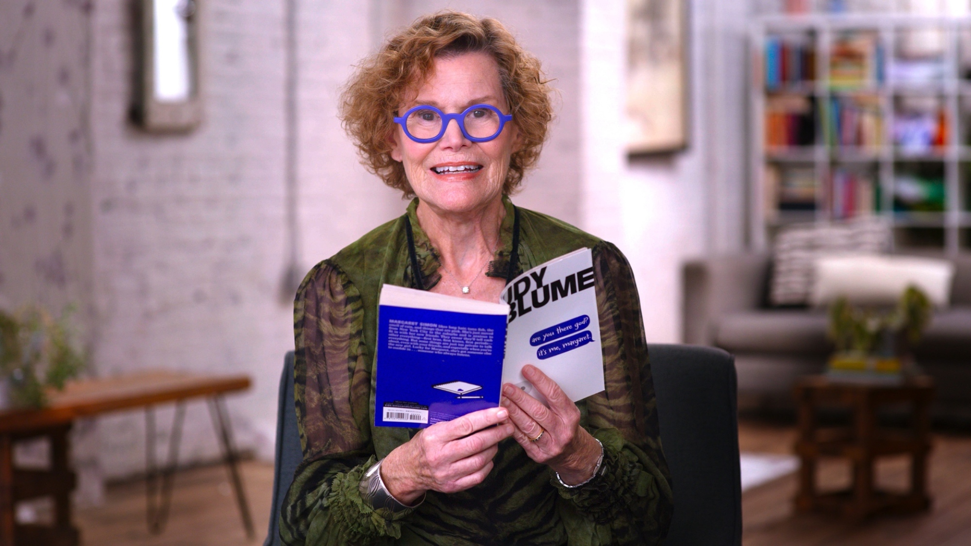 Une femme aux cheveux bouclés courts et aux lunettes bleu vif tient un exemplaire d'un livre intitulé "Es-tu là, mon Dieu ?  C'est moi, Marguerite."