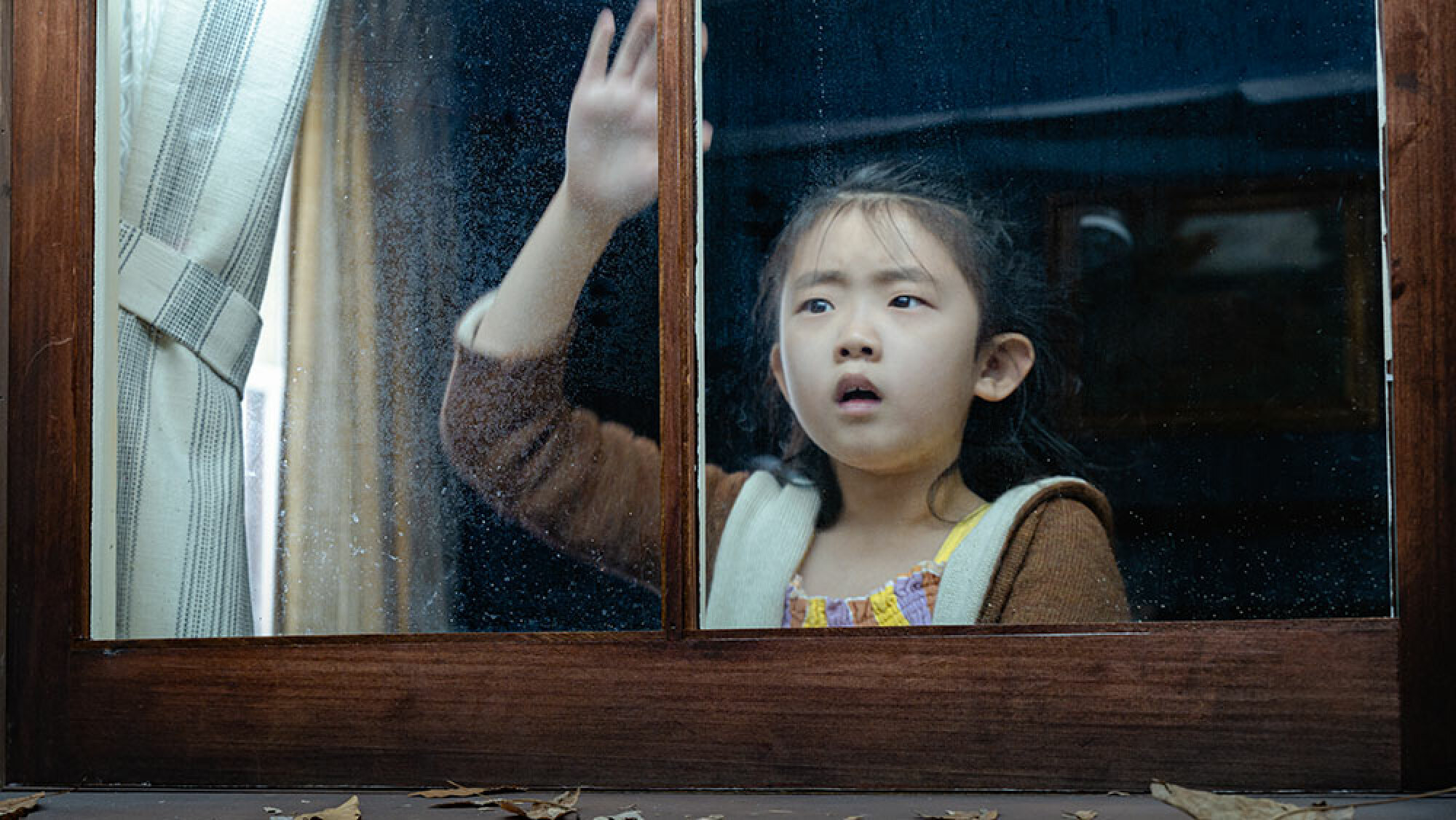Une petite fille regarde par la fenêtre d'une cabane, l'air effrayée.