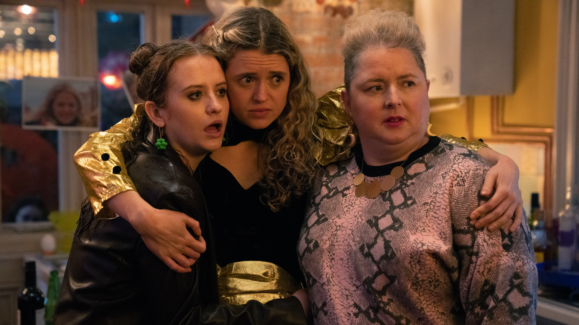 Trois femmes se tiennent dans un appartement au milieu d'une fête, l'air choquée.
