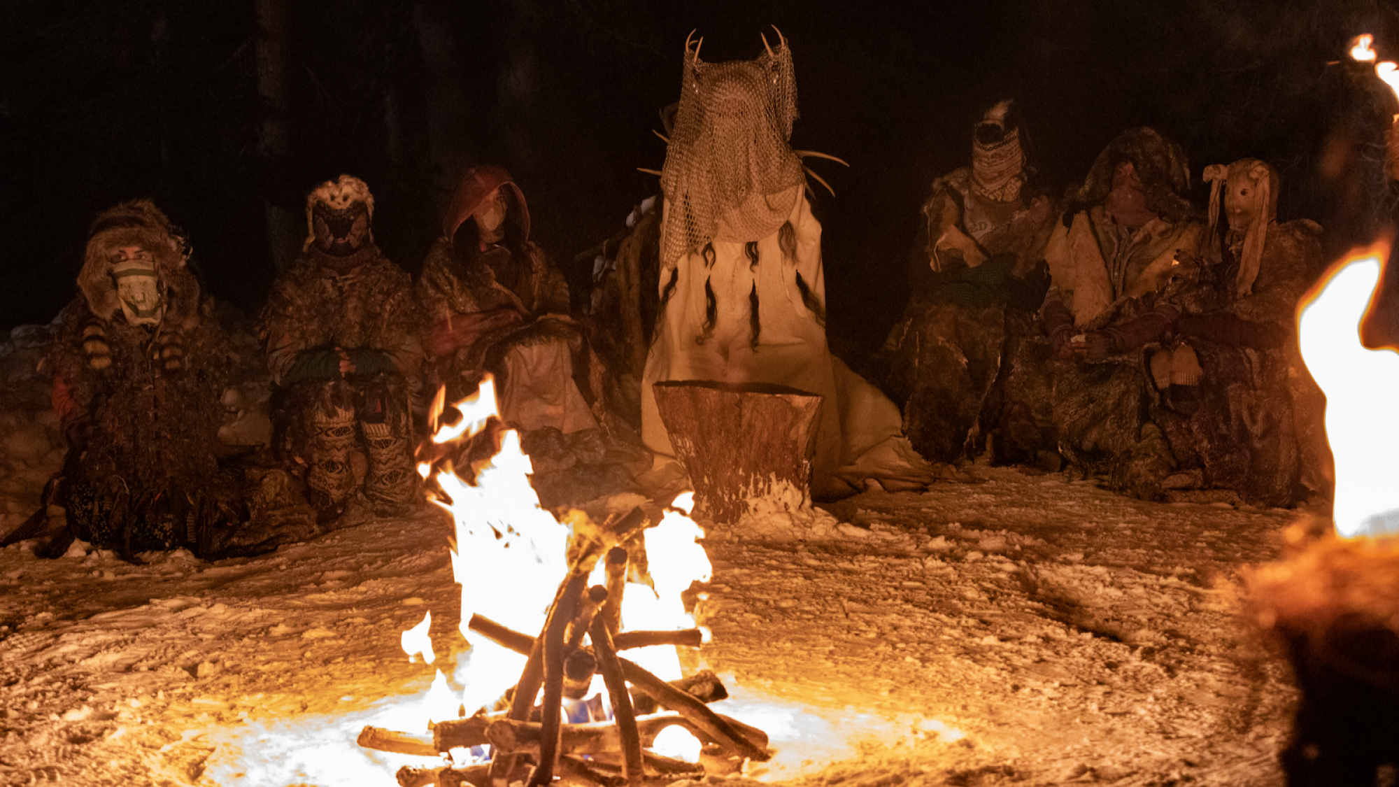 Un groupe portant des masques d'animaux et des cornes est assis autour d'un feu. 