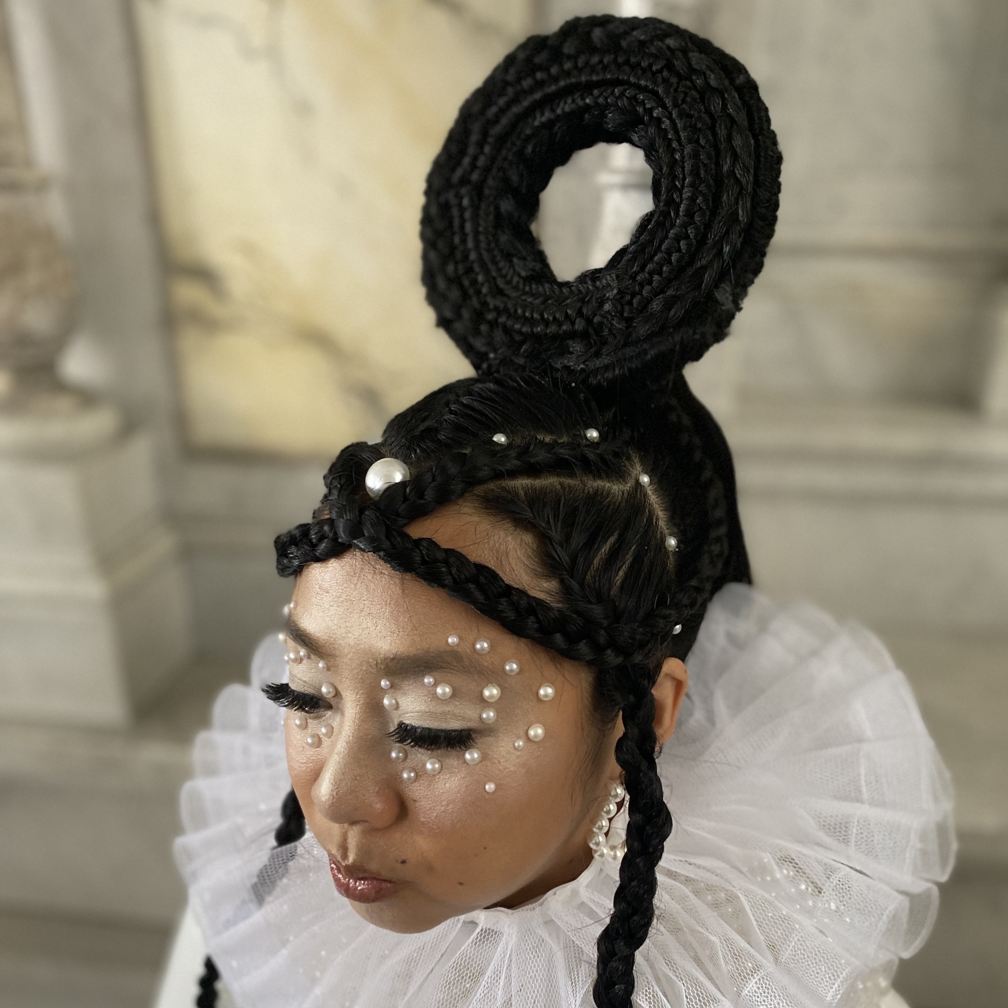 Une femme avec un maquillage blanc chatoyant et des perles sur son visage.  Un bagel fait de cheveux tressés est posé sur le dessus de sa tête.