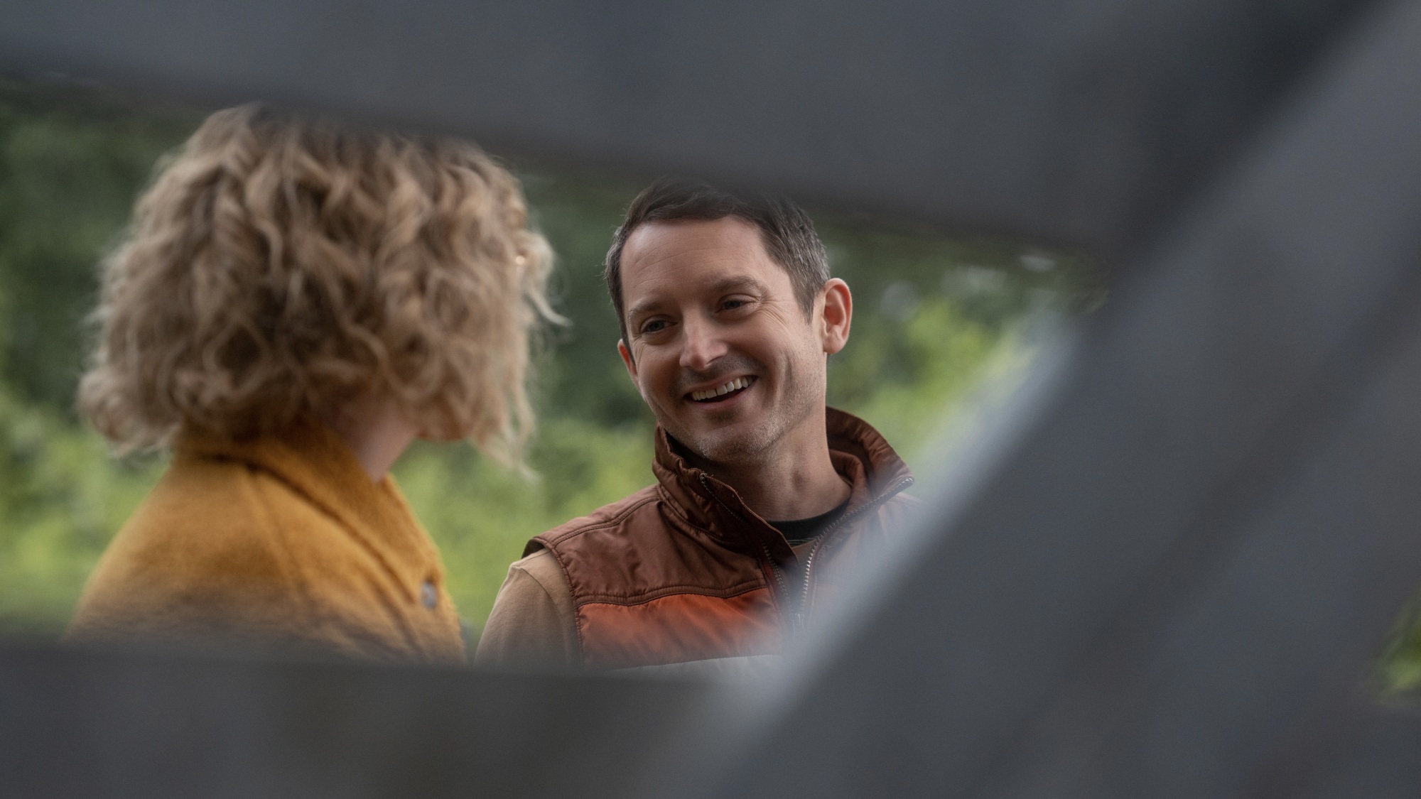 Une femme en manteau jaune et un homme souriant en gilet marron et orange regardent à travers les lattes d'une clôture.
