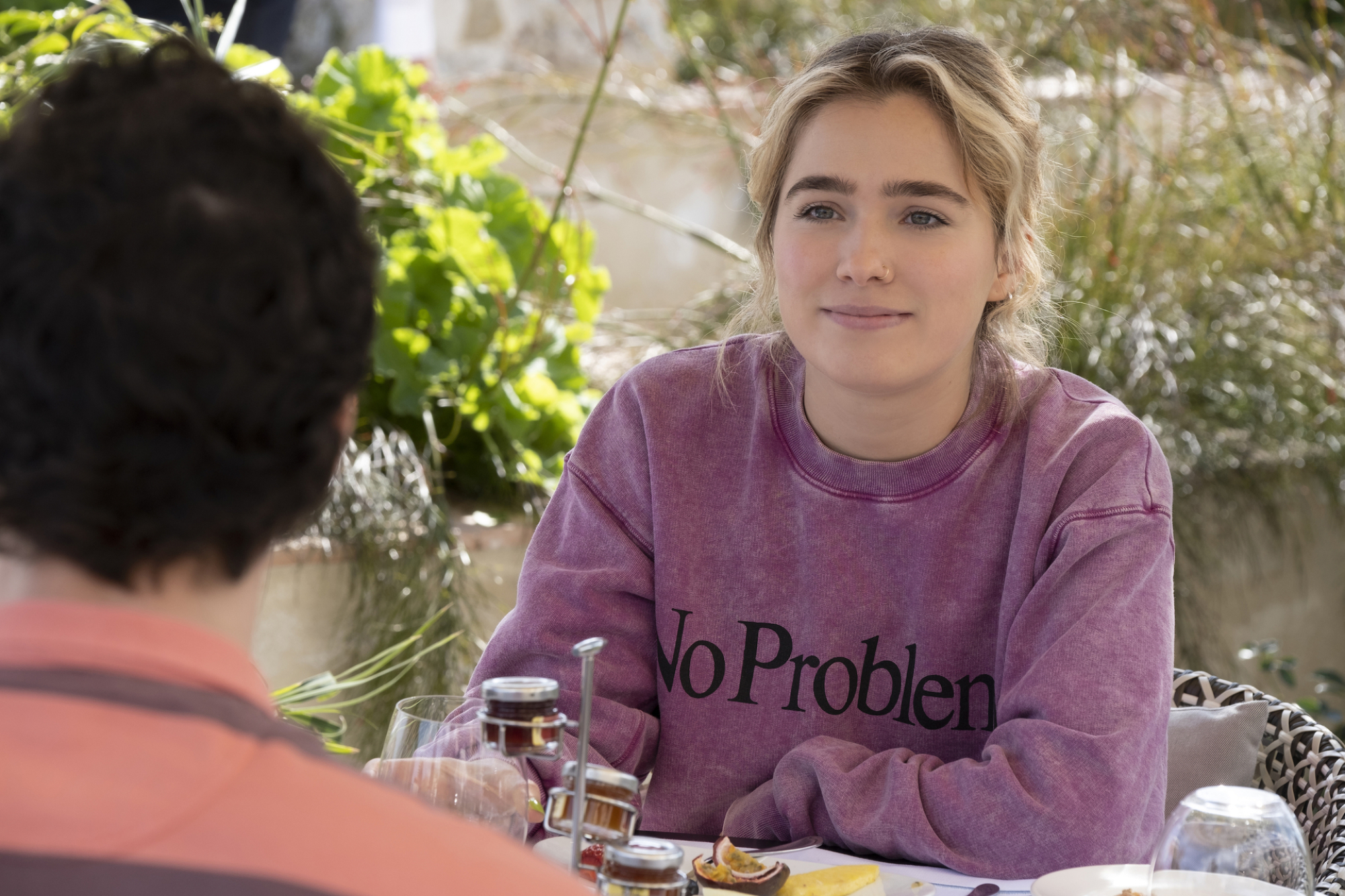 Portia est assise à une table de petit-déjeuner et porte un sweat-shirt violet délavé à l'acide sur lequel on peut lire : 