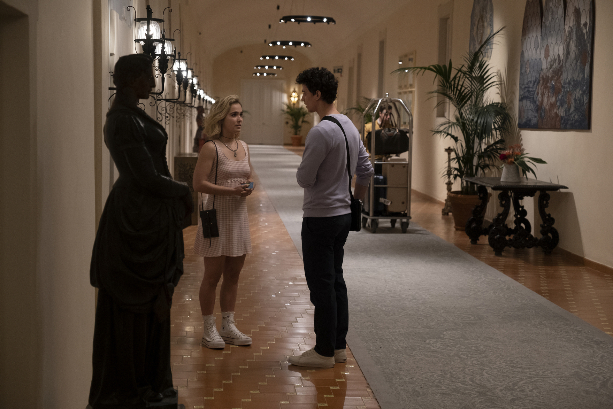 Portia et Albie discutent dans le couloir de l'hôtel.  Portia porte une robe à carreaux rose. 