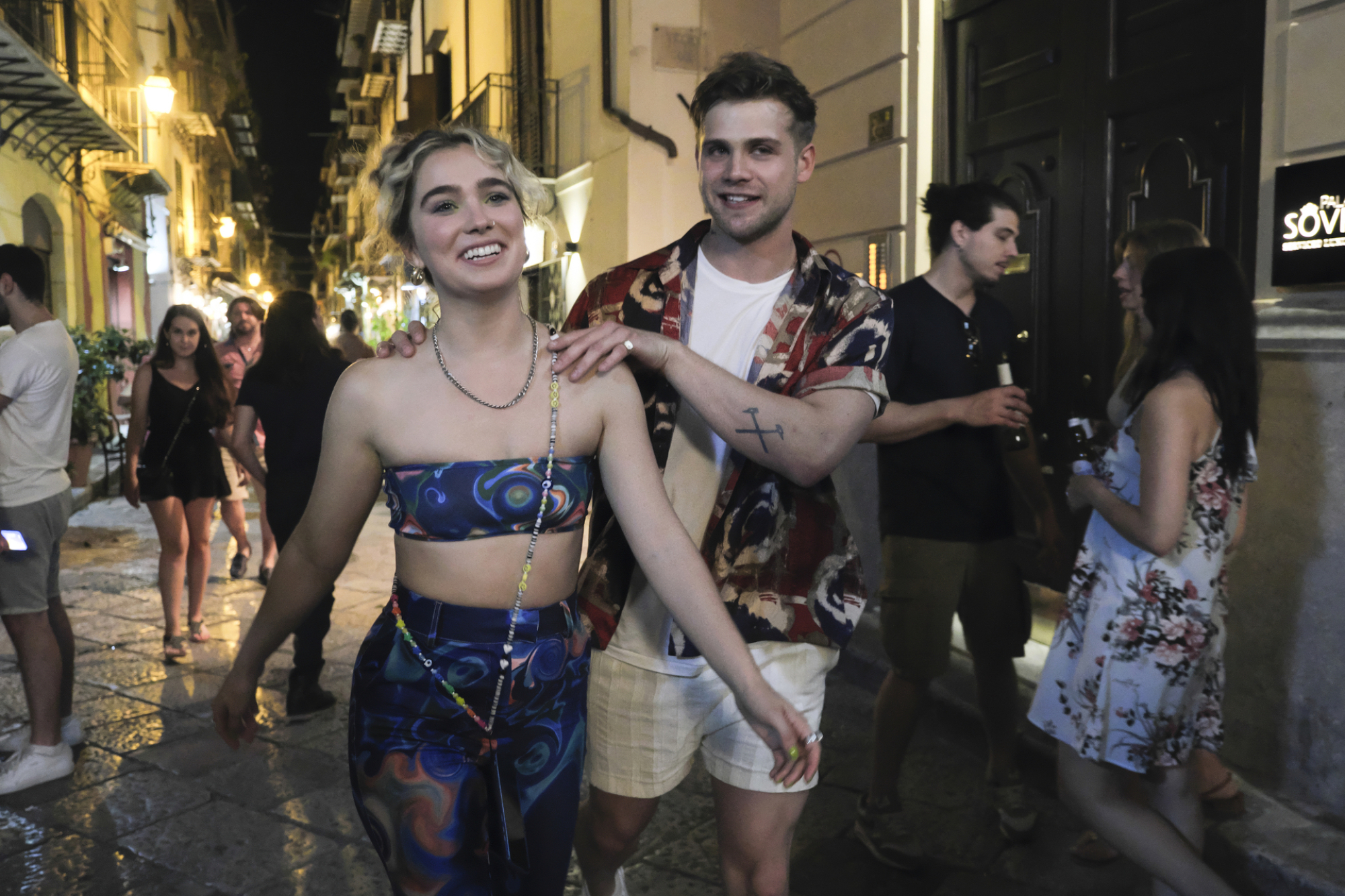 Jack a ses mains sur les épaules de Portia alors qu'ils marchent dans une rue de Palerme la nuit.  Portia porte un ensemble bleu assorti. 