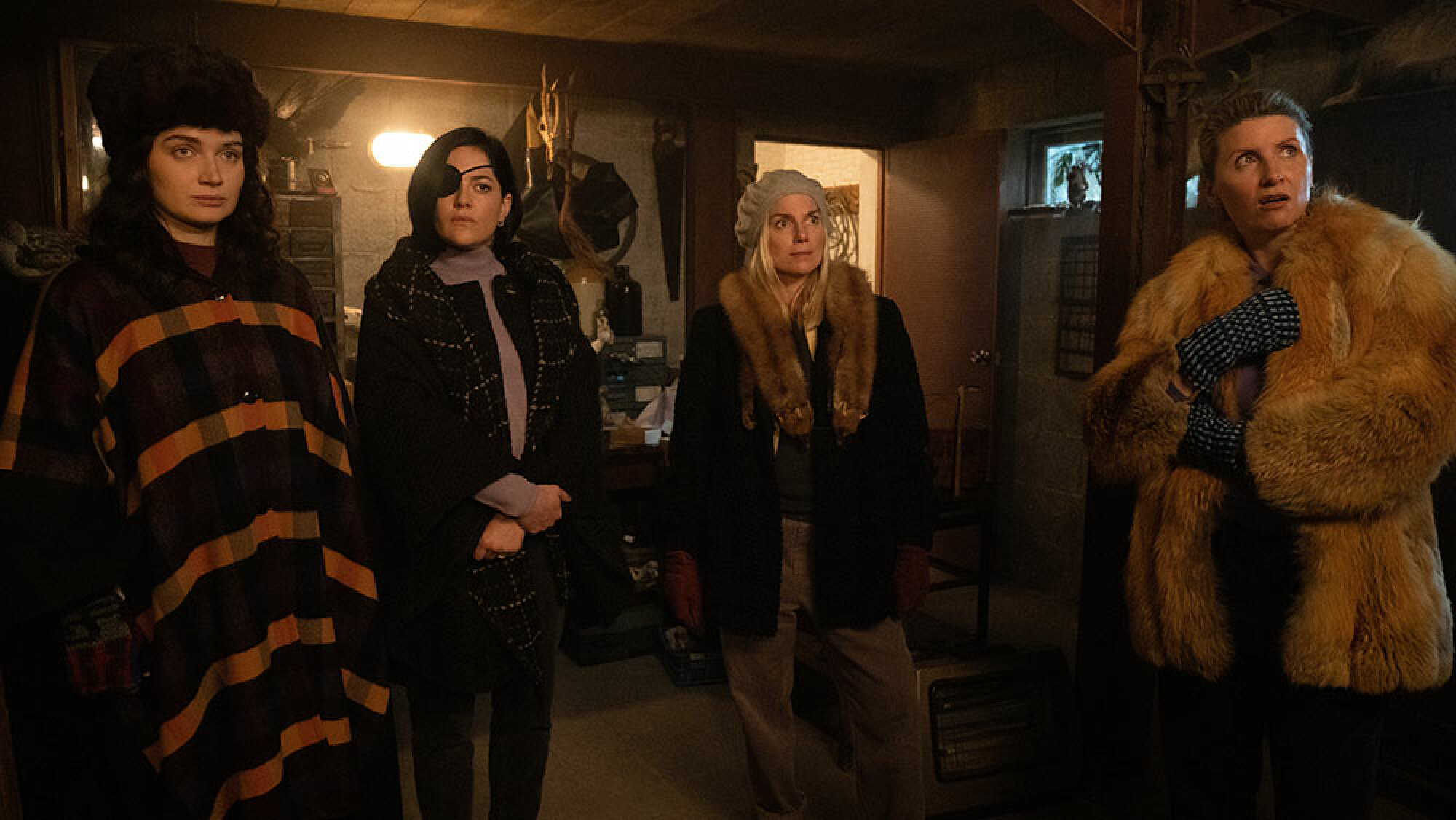 Quatre femmes se tiennent en manteaux dans le sous-sol sombre d'une maison.