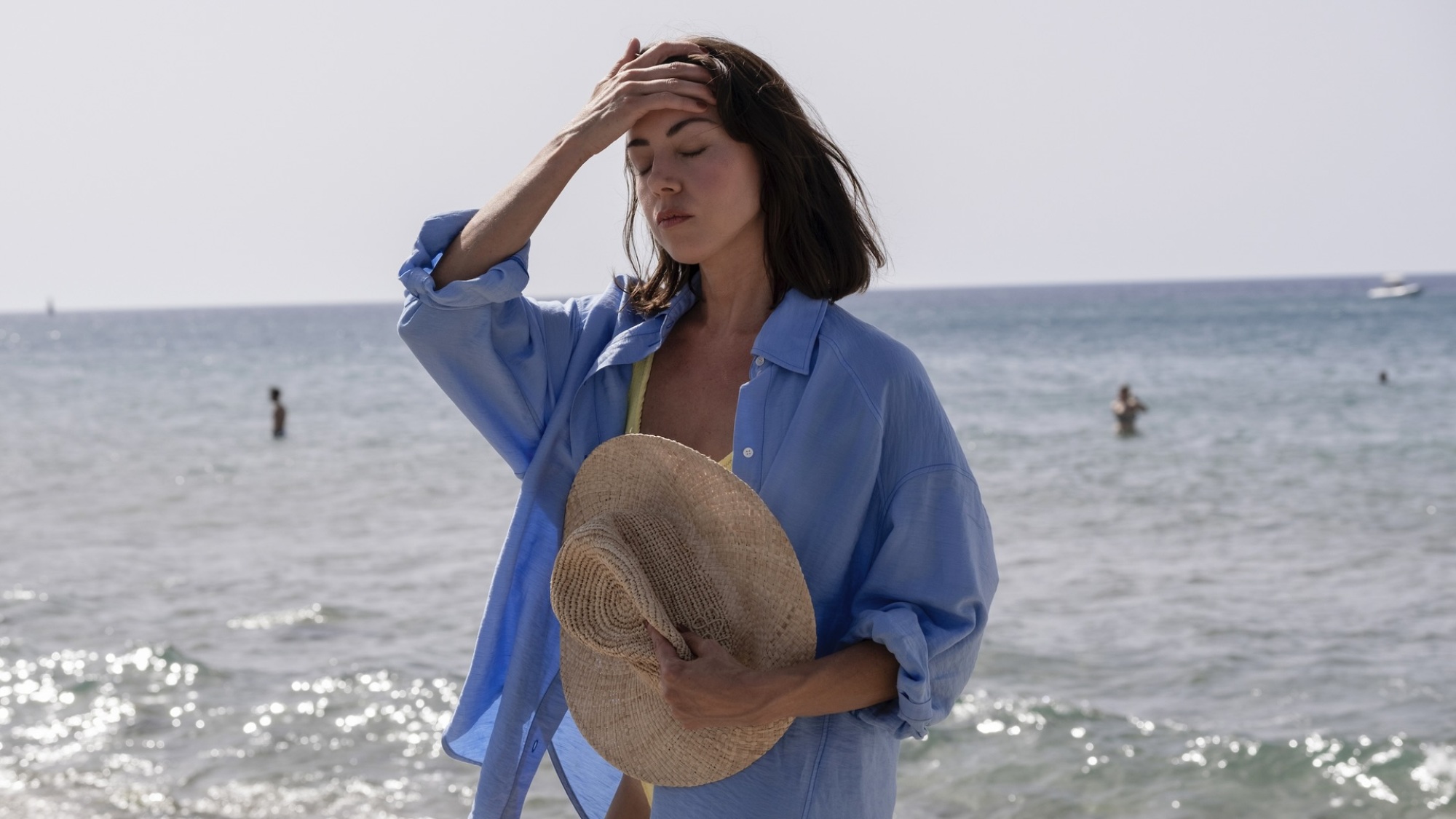 Une femme en chemise bleue se tient la main sur le front avec l'océan derrière elle.