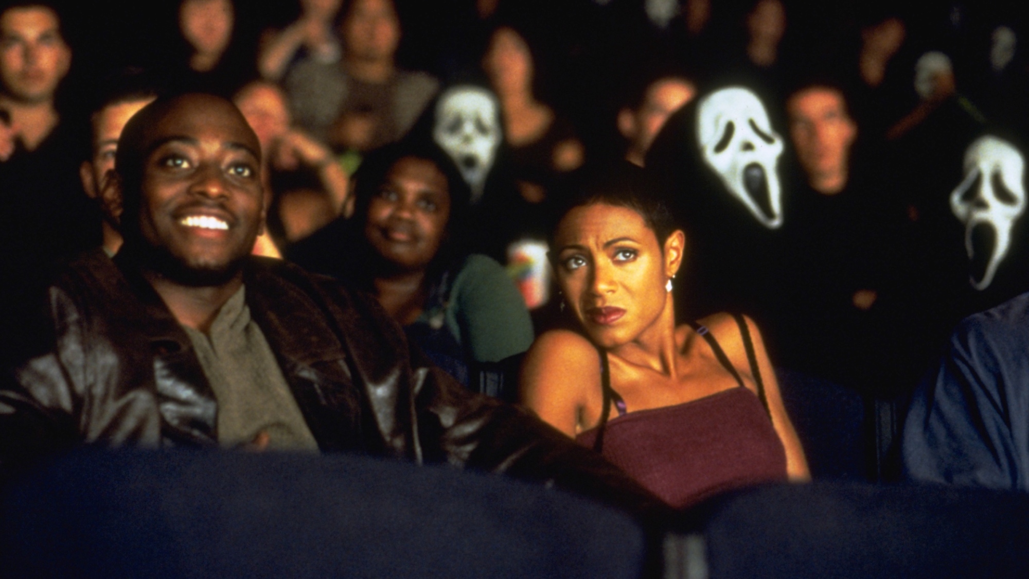 Un homme et une femme à un rendez-vous sont assis ensemble dans une salle de cinéma, entourés de personnes portant des masques Ghostface.  L'homme sourit et la femme a l'air effrayée.