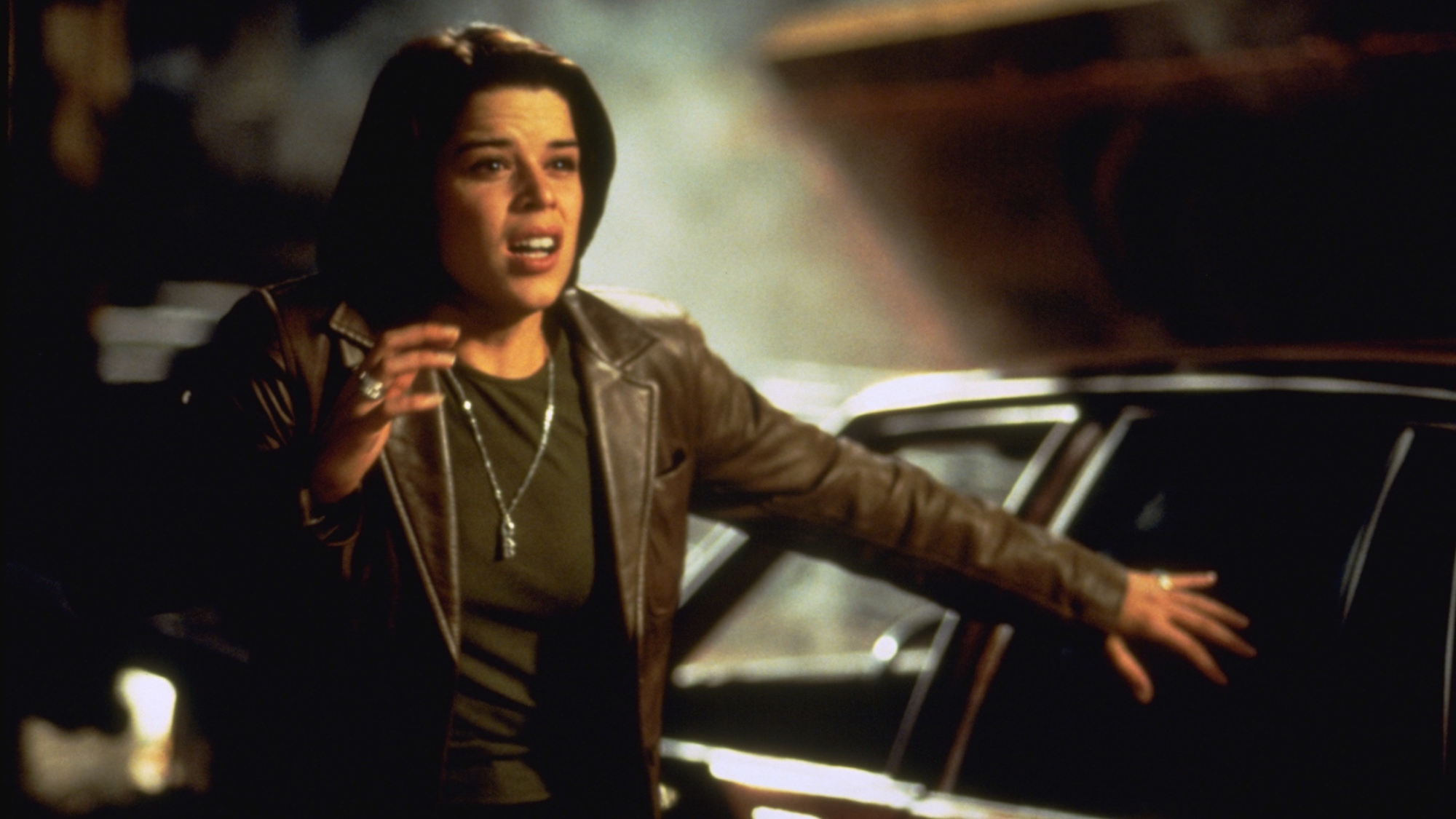 Une femme brune se tient près d'une voiture, tenant une main près de son visage, qui a l'air effrayé.