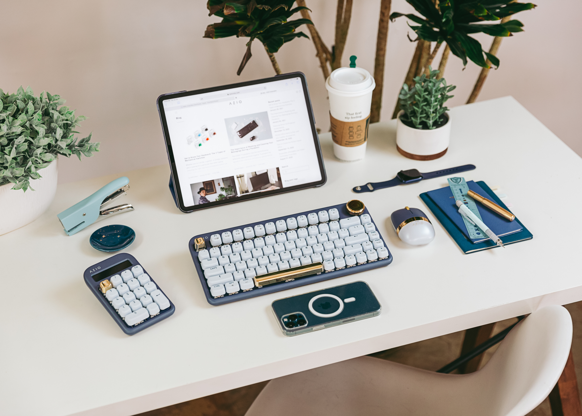 Une configuration de travail sur un bureau blanc composé d'un iPad et d'un clavier bleu foncé avec des accents dorés, calculatrice assortie.  souris assortie et un iPhone face vers le bas.