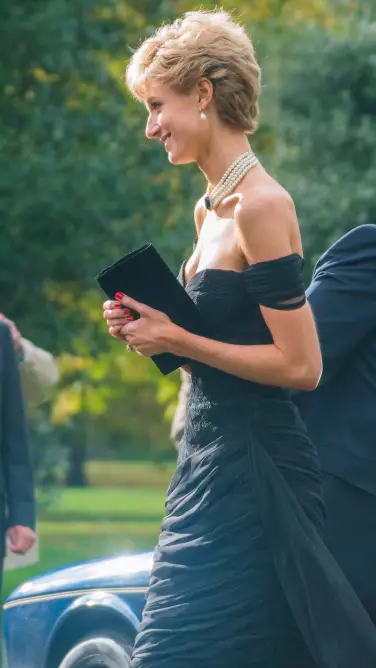 Une femme vêtue d'une robe de soirée noire à épaules dénudées se promène en tenant une pochette.