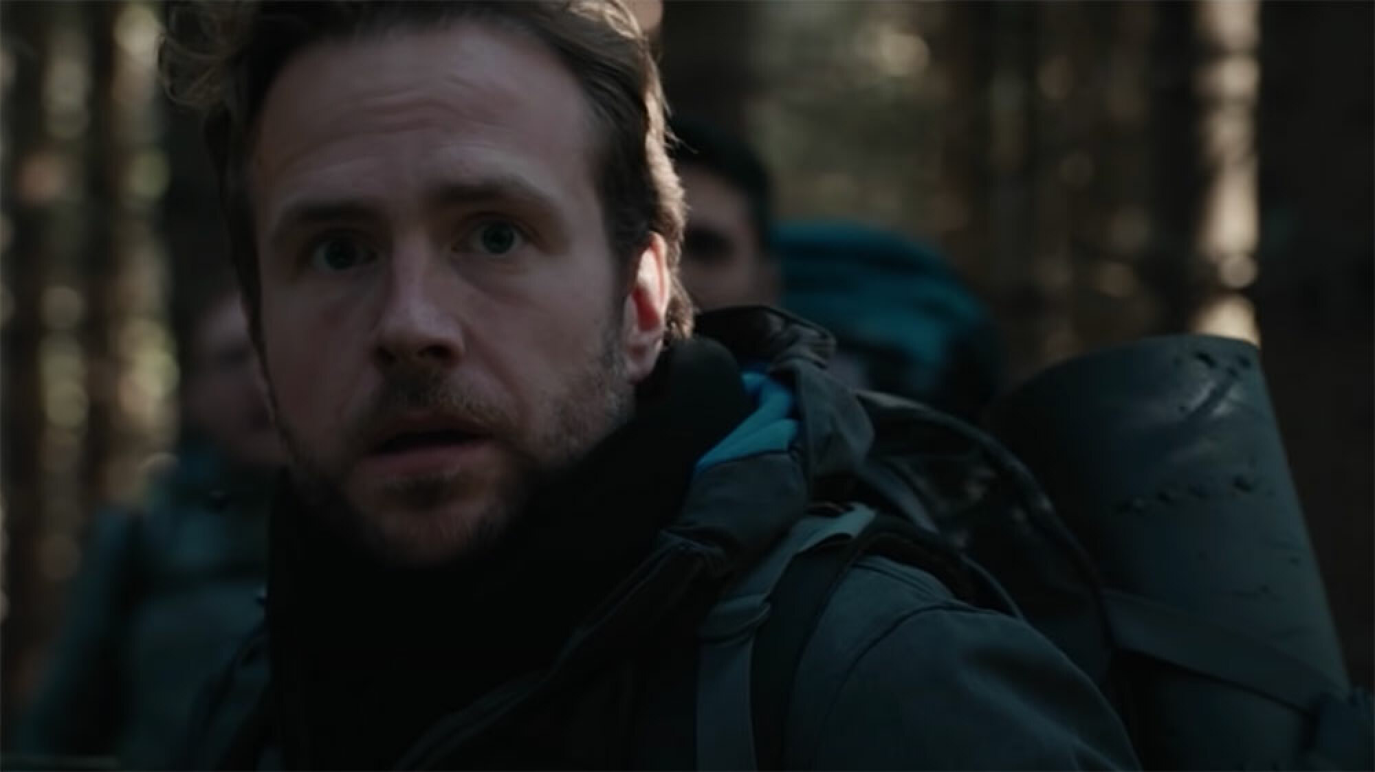 Un homme avec un sac à dos dans les bois regarde dans la direction de la caméra, l'air effrayé.