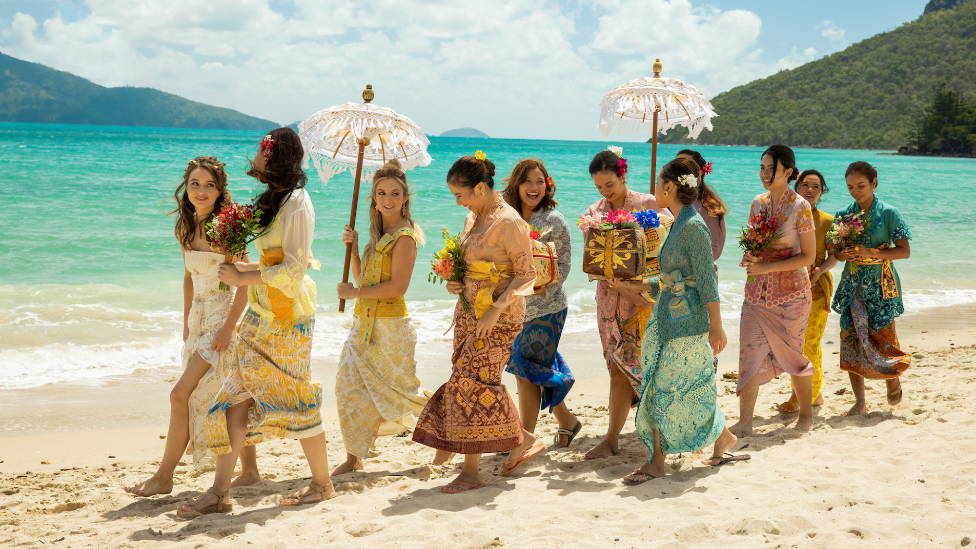 Une procession de femmes portant diverses tenues de mariage balinaises marche le long d'une plage.