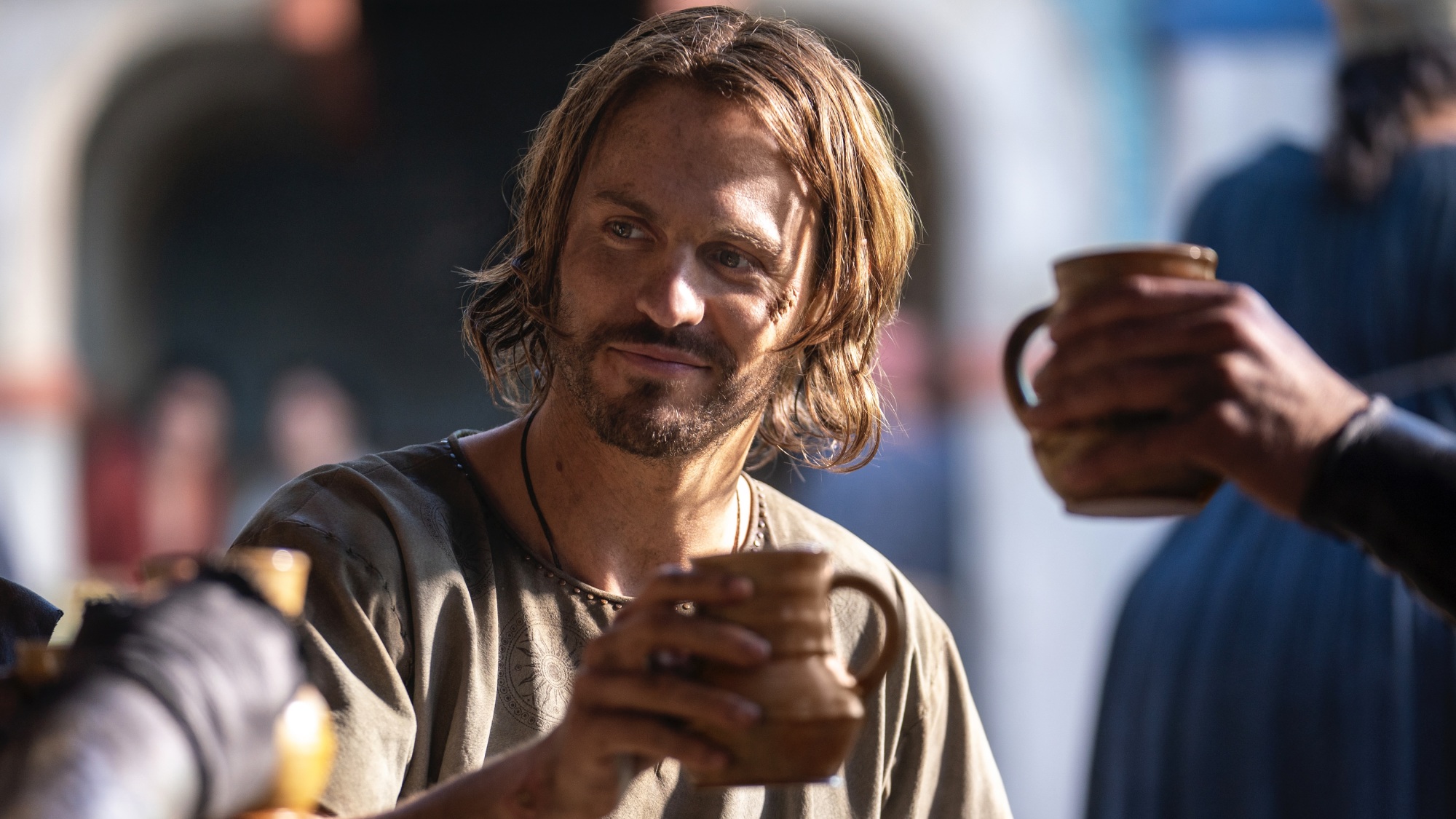 Un homme aux cheveux bruns mi-longs lève une tasse en céramique brune pour porter un toast.