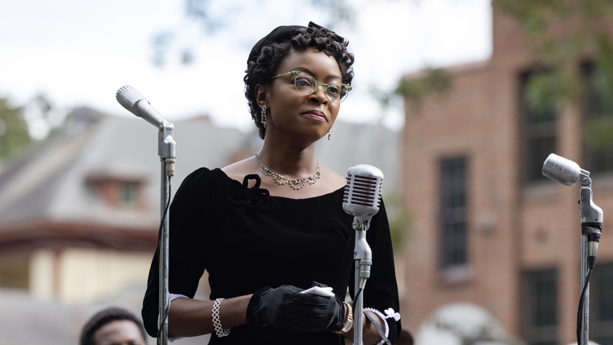 Une femme vêtue d'une robe noire et d'un chapeau se tient devant plusieurs microphones, prononçant un discours.