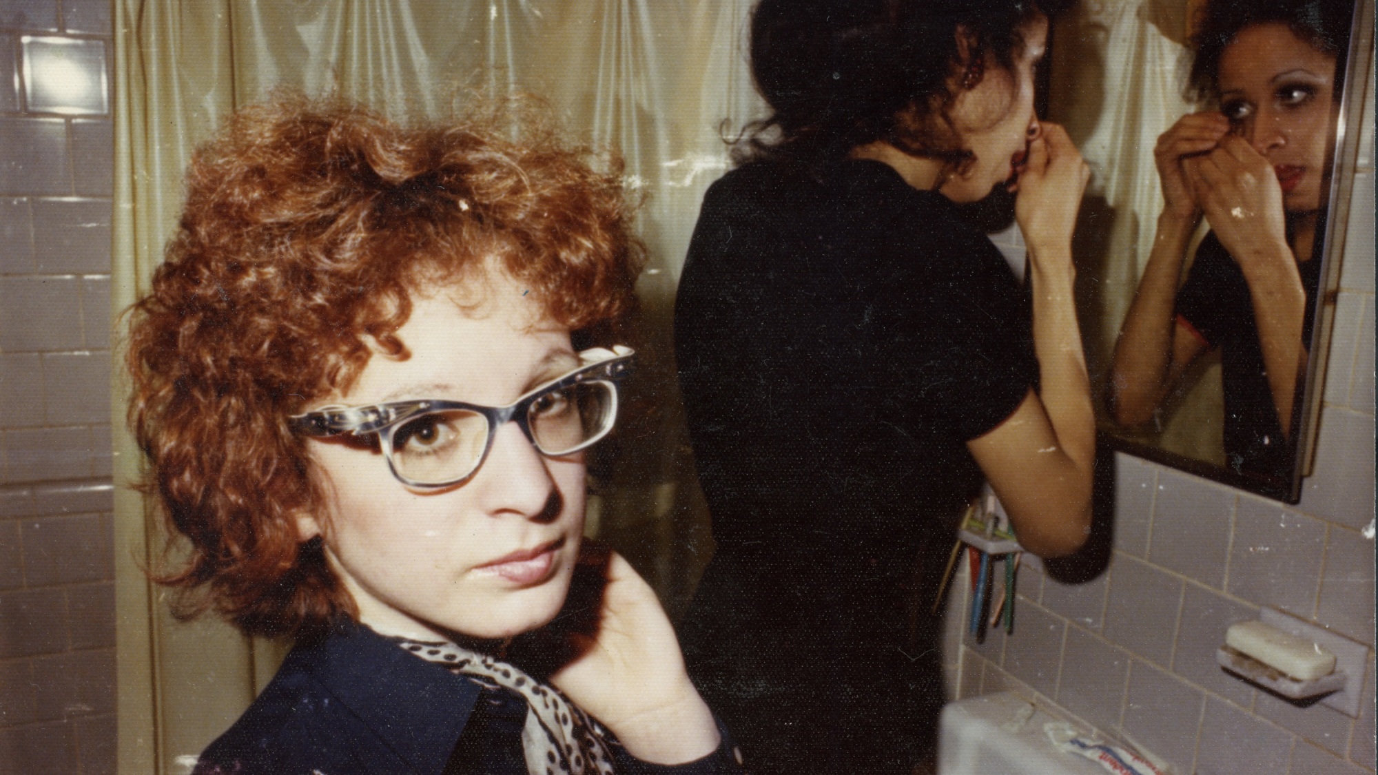 Deux femmes dans une salle de bain : l'une d'elles, aux cheveux roux bouclés et à lunettes, regarde directement dans l'appareil photo, tandis que l'autre se maquille dans le miroir.