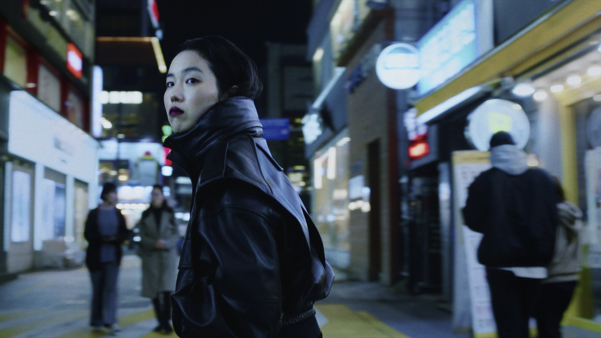 Une femme en manteau noir à col haut et rouge à lèvres dans une rue de Séoul la nuit.