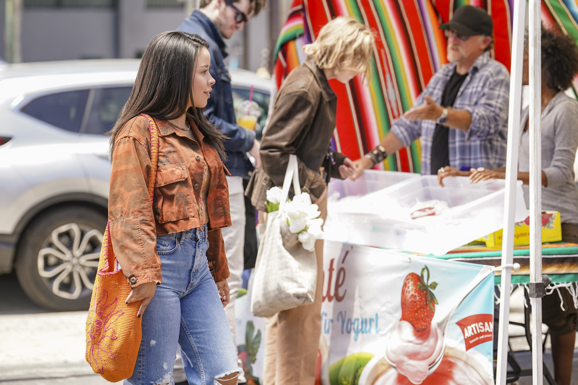 Une jeune femme vêtue d'une veste et d'un sac orange passe devant un étal de marché de producteurs.