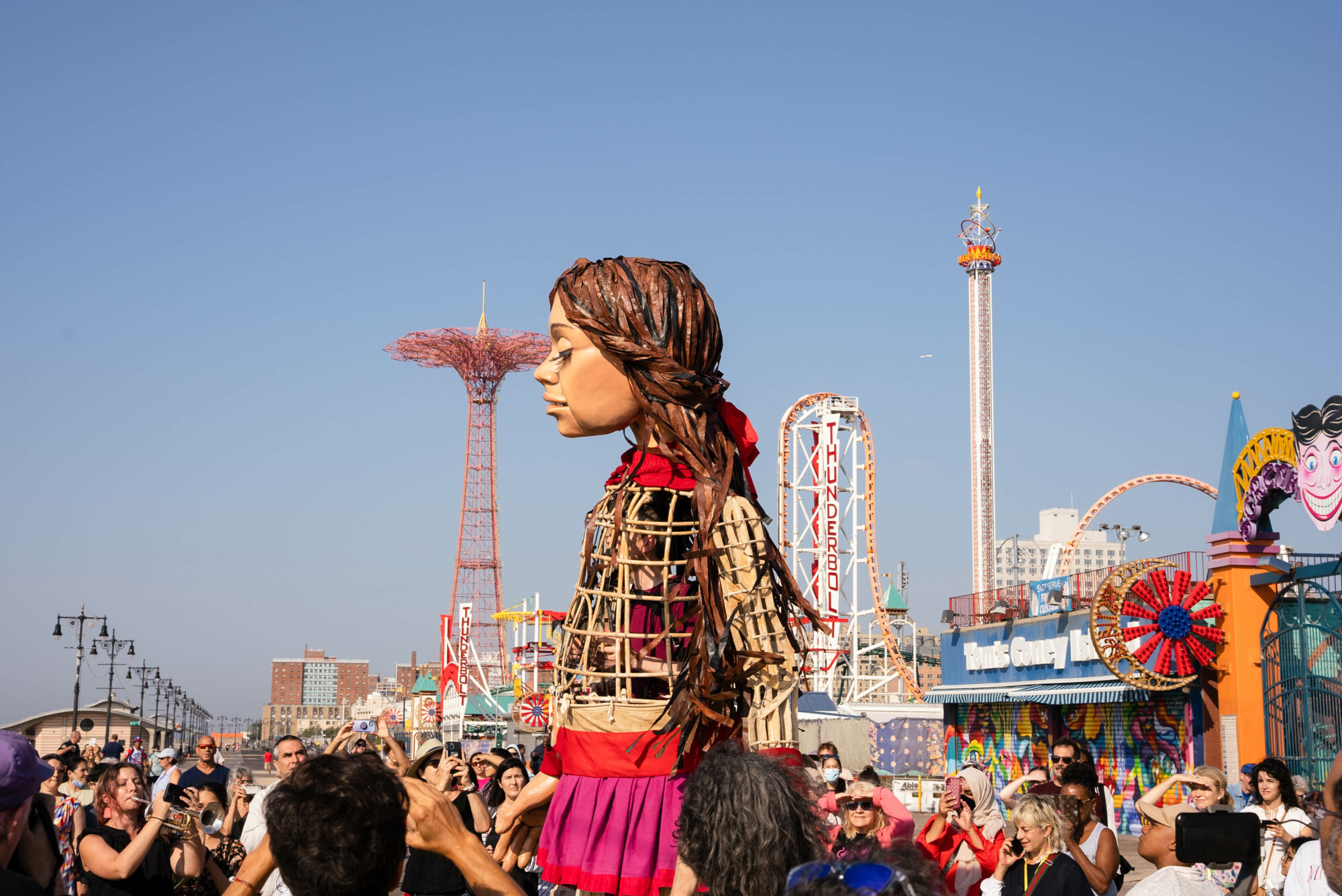 Amal se tient de profil sur la promenade de Coney Island.  Une foule l'entoure et vous pouvez voir des manèges en arrière-plan.