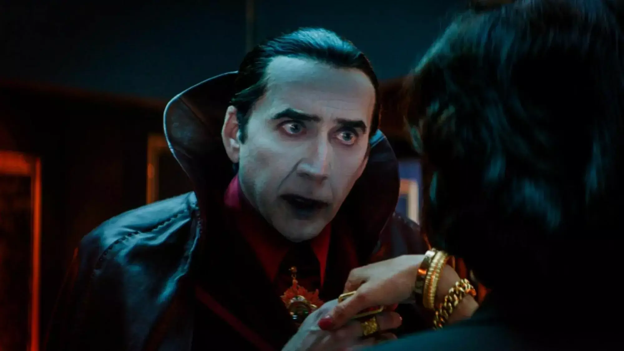 Nicolas Cage incarne Dracula dans "Renfield," avec les cheveux lissés en arrière, un visage peint en blanc et des lèvres rouges, serrant la main d'une femme.