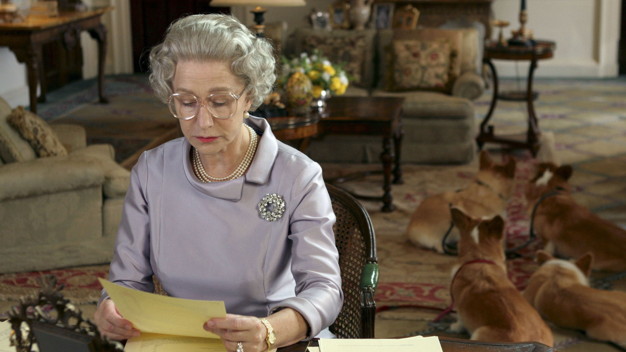 Un acteur déguisé en reine Elizabeth II lisant des lettres avec ses corgis en arrière-plan.