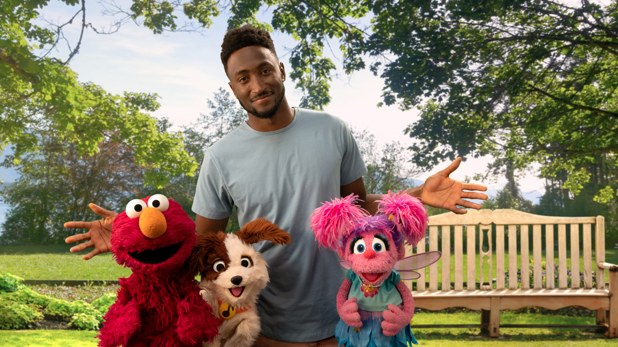 Marques Brownlee, Elmo, Abby et Tango le chien se tiennent devant un banc dans un parc ensoleillé.