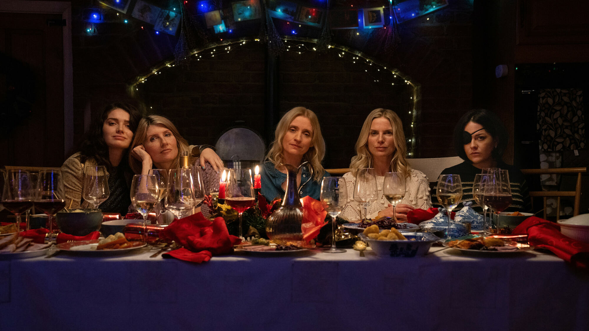 Cinq femmes s'assoient à une table à manger festive en regardant la caméra d'un air absent.