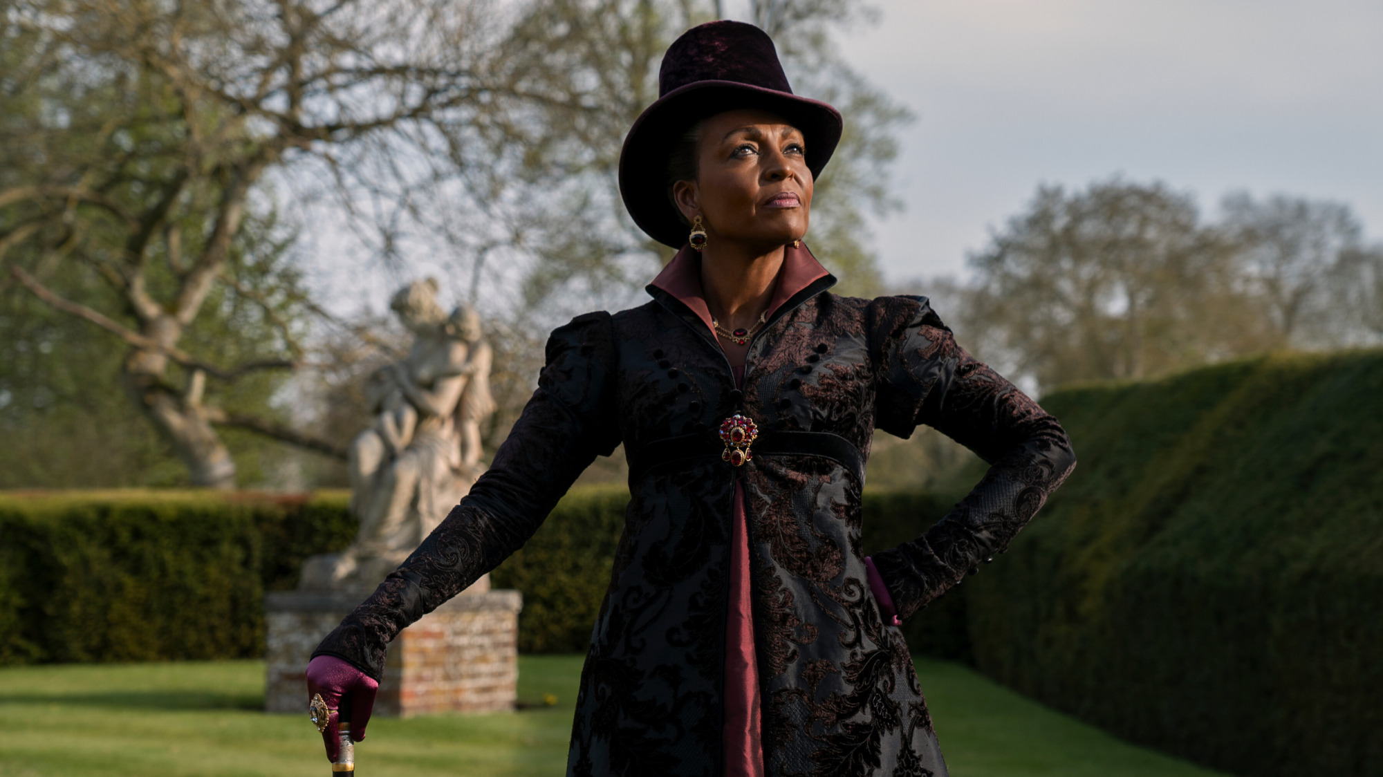 Une femme portant un manteau noir et un chapeau se tient dans un jardin. 