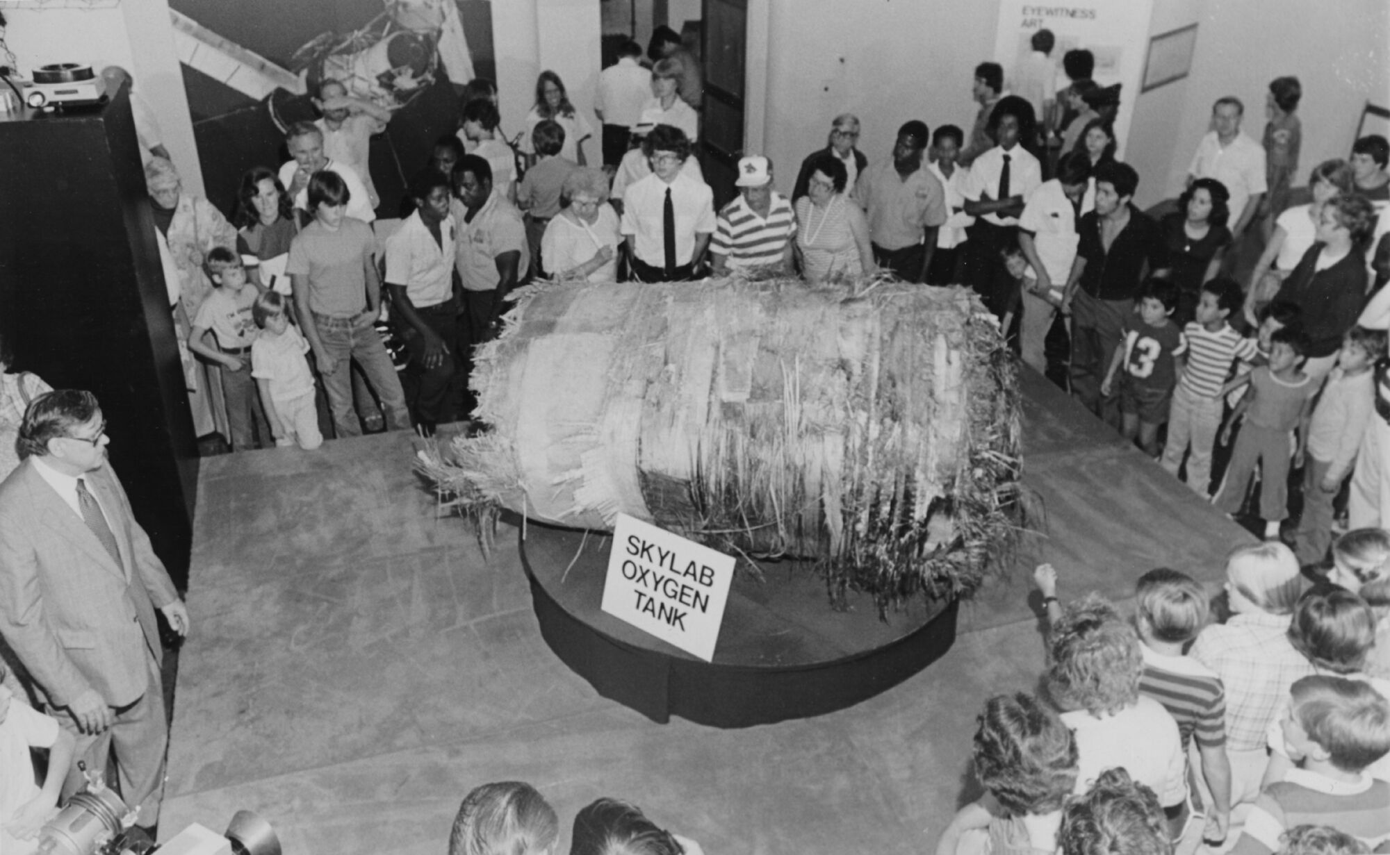 Une photo en noir et blanc de 1979. Un groupe de visiteurs du musée se rassemble en cercle autour d'un gros morceau de métal déchiqueté en forme de cylindre.  Il repose sur un piédestal rond avec un panneau indiquant 