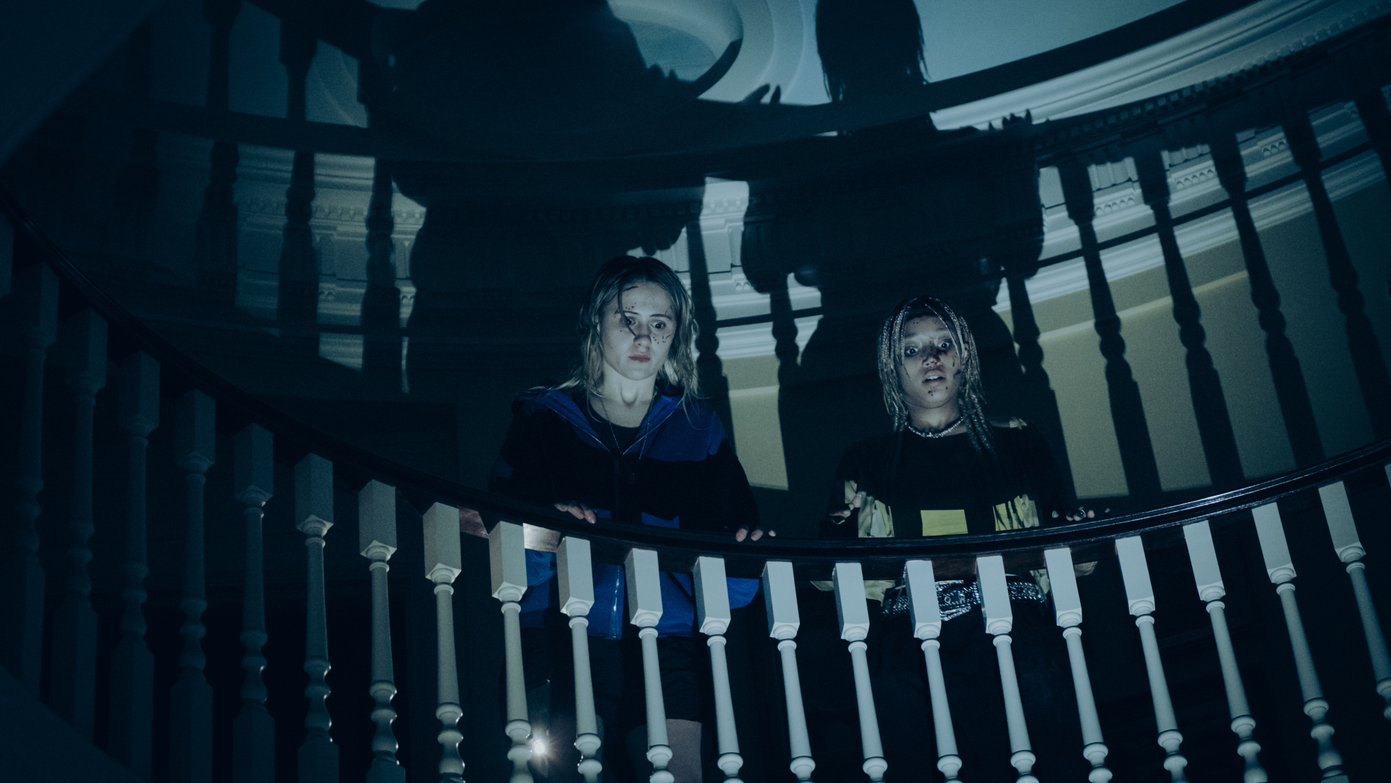 Deux jeunes femmes tachées de sang se tiennent en haut d'un escalier et regardent vers le bas.